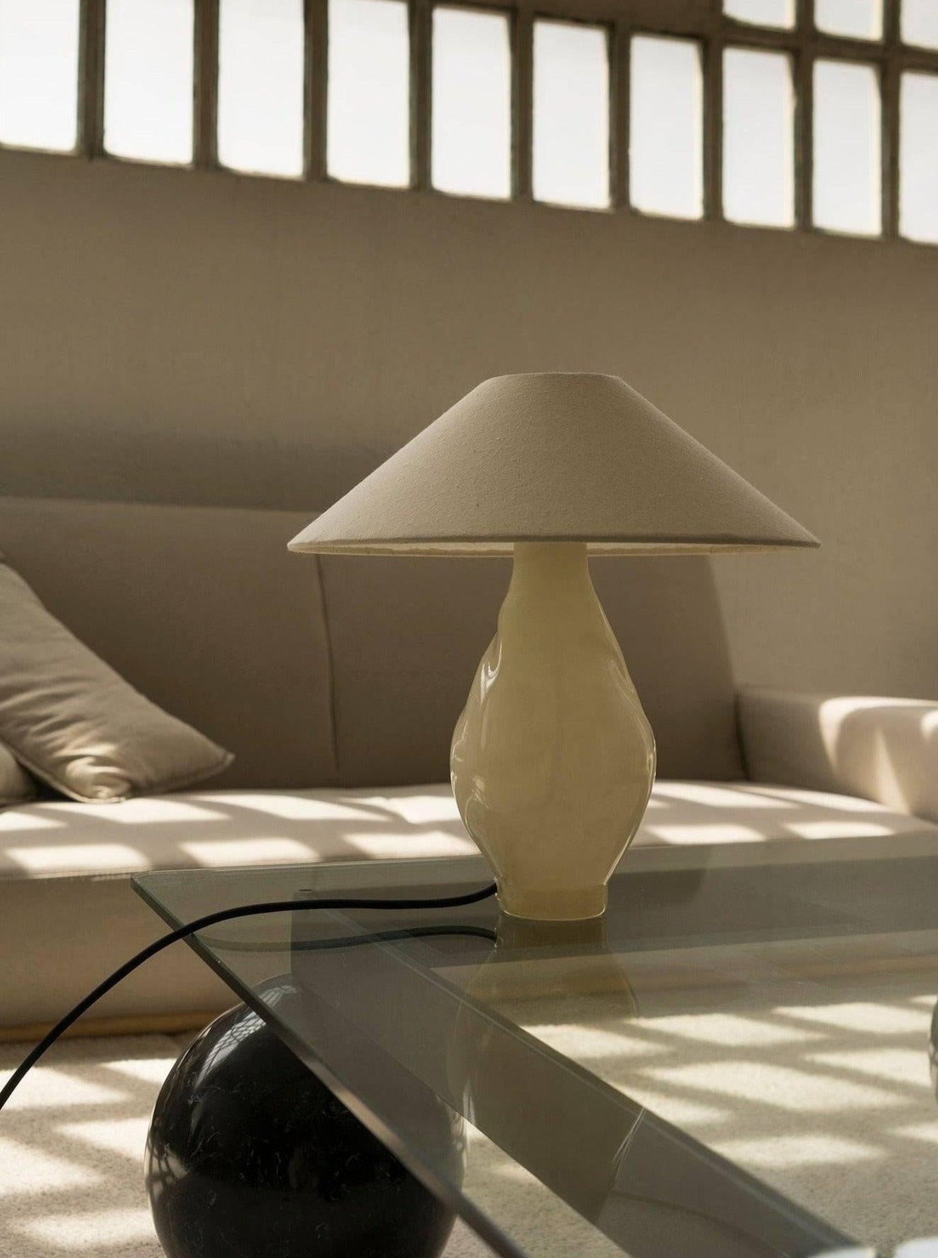 A minimalist living room with a beige sofa and cushions embodies contemporary design. The glass coffee table is adorned by the Los Objetos Decorativos Beige Conical Glass Lamp, while sunlight through a grid-like window casts soft shadows to enhance the aesthetic.