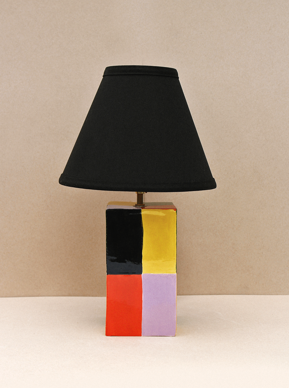 Table lamp with a multicolored ceramic base featuring bold blocks of black, red, gold, and purple, topped with a simple black lampshade, against a beige background. The Milagro Lamp (Chico) by Casa Veronica is UL listed.