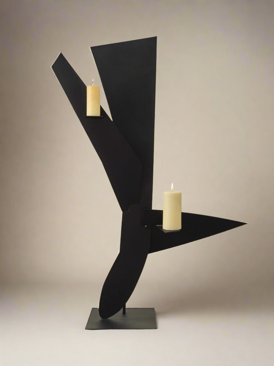 The Bird Candleholder by Animate Objects features a modern, abstract black powder coat finish with angular, geometric shapes. Two pillar candles rest on horizontal projections above a flat rectangular base, all set against a neutral background.