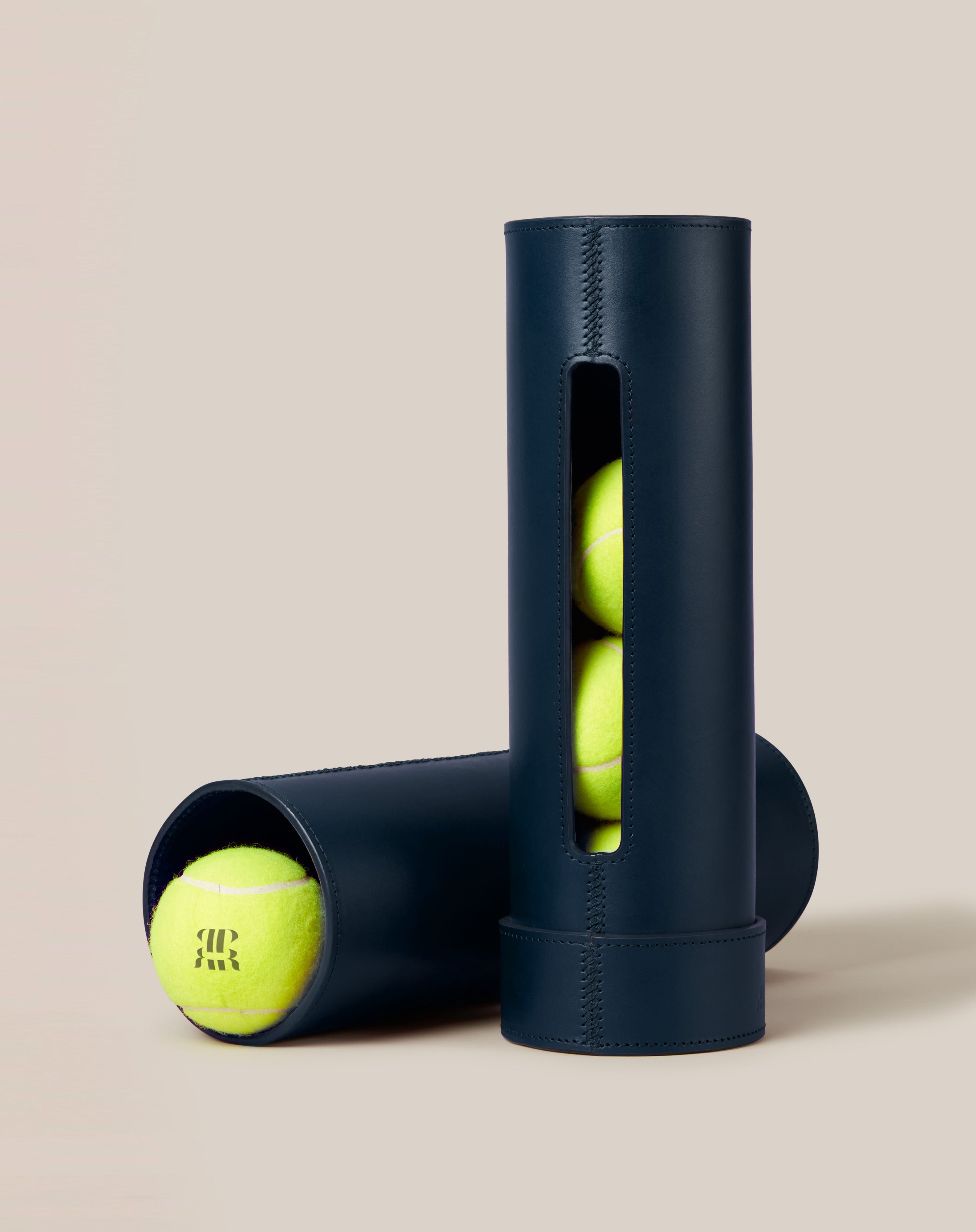 Two elegant, dark blue cylindrical Leather Tennis Ball Holders from Paradise Row are crafted from sustainable vegetable-tanned leather and contain bright yellow tennis balls. One holder stands upright with a side opening revealing three balls, while the other rests horizontally with one ball partially visible. The background is a neutral beige.