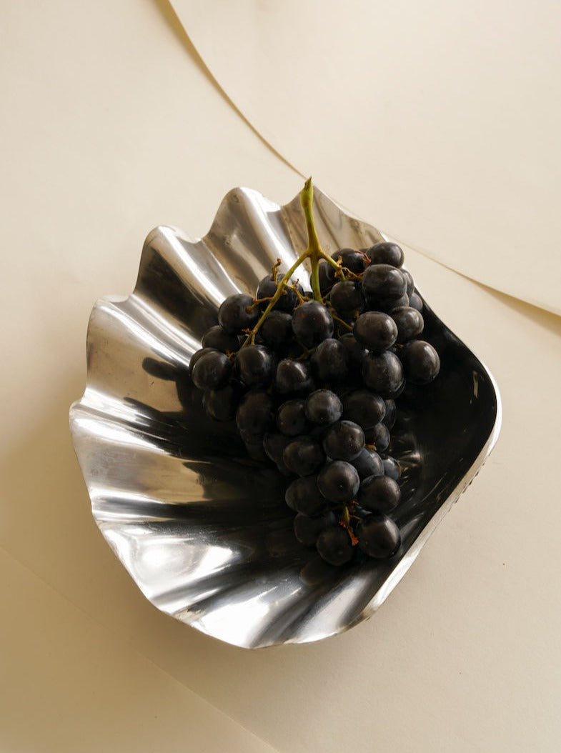 A bunch of dark grapes is placed in a shiny, silver, Shell-Shaped Fruit Bowl by Les Objets Amsterdam resting on a beige surface. The composition has a minimalistic and artistic feel.