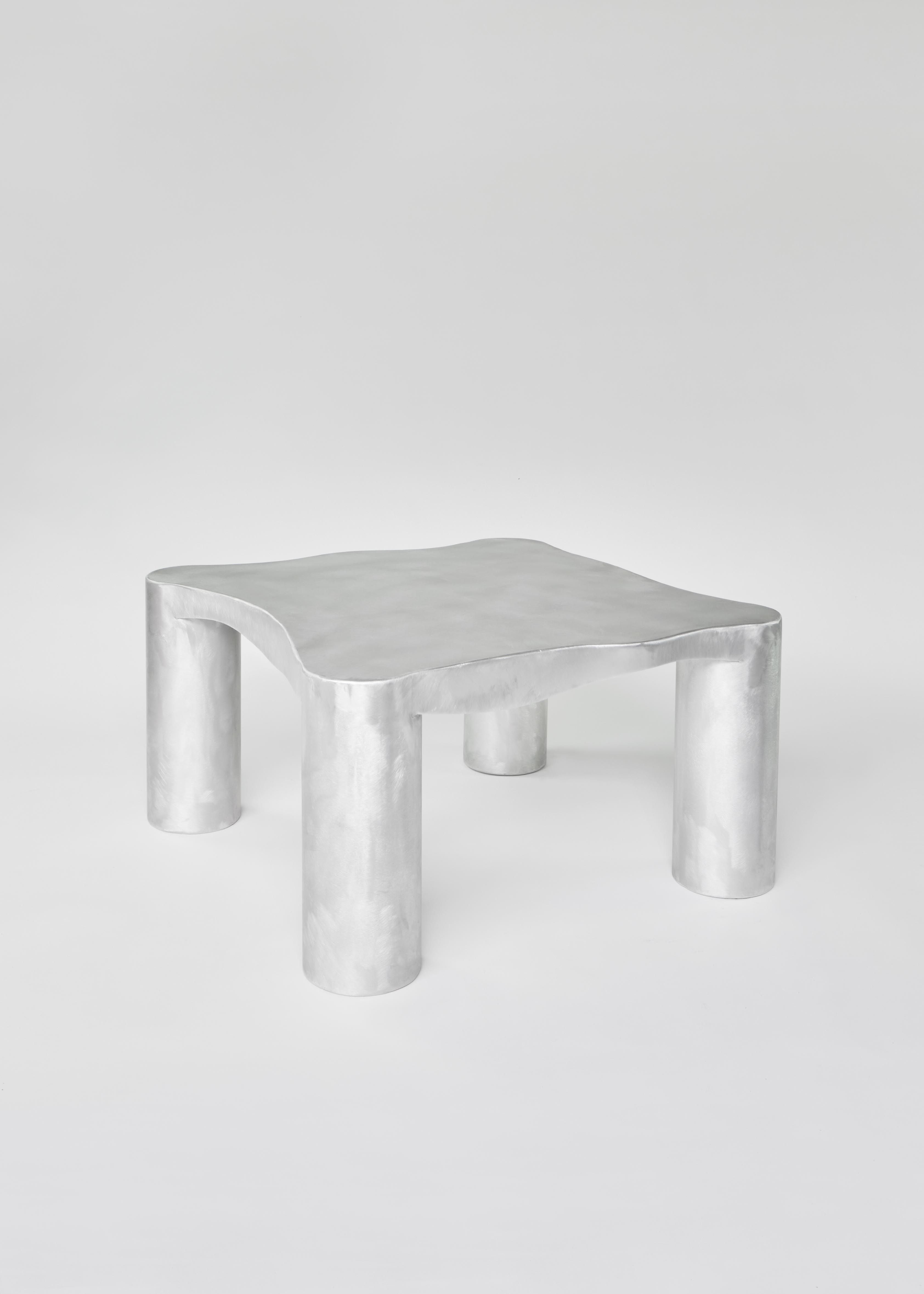 The Coffee Table ‘Not in Service’ by Six Dots Design is a minimalist masterpiece, featuring a handcrafted aluminium design with four sturdy cylindrical legs and an irregularly shaped top. Its sleek, modern aesthetic is complemented by a smooth, brushed metal finish, making it an ideal addition to both indoor and outdoor spaces against a plain white background.