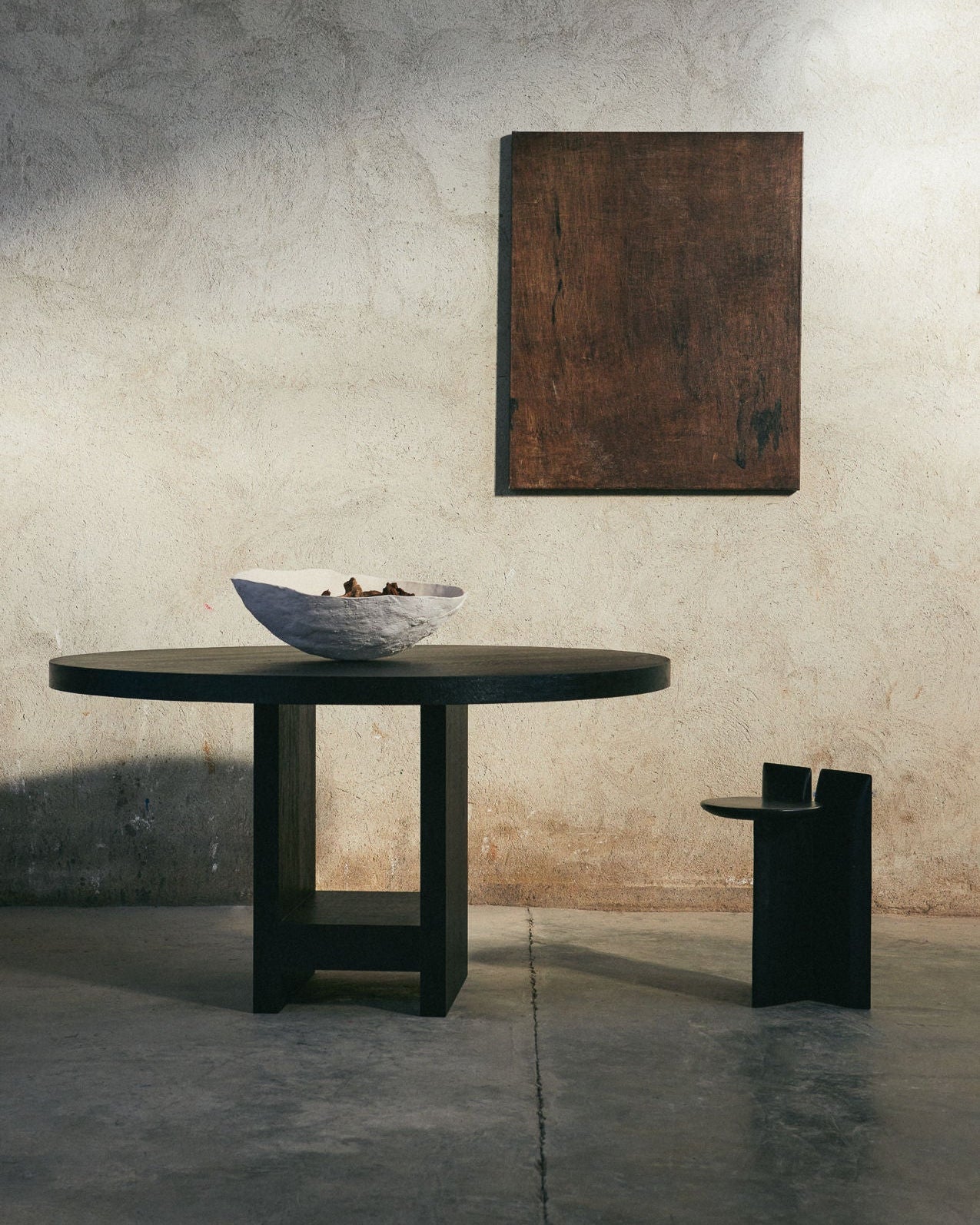 A minimalist room features the Sister Table by ASERIES OF OBSTACLES, SL, with a large light-colored bowl on top, paired with a simple wooden chair and abstract artwork on a textured beige wall. The concrete floor and soft natural light enhance the elegance of natural materials.