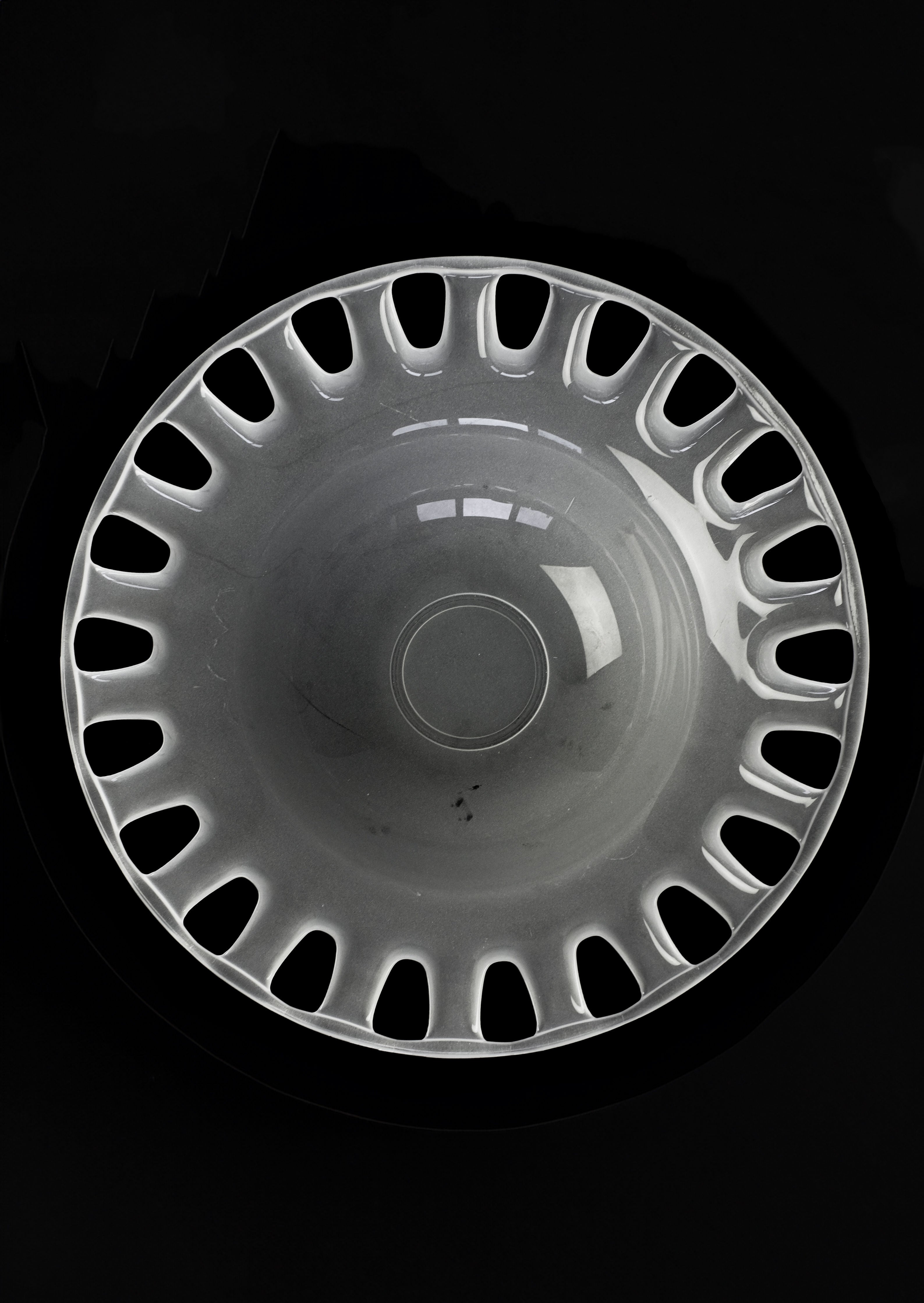 A symmetrical, round hubcap with a glossy surface and cut-out holes along the outer edge resembles the elegance of septembre studios' Large Bowl of Milk Glass, set against a sleek black background.