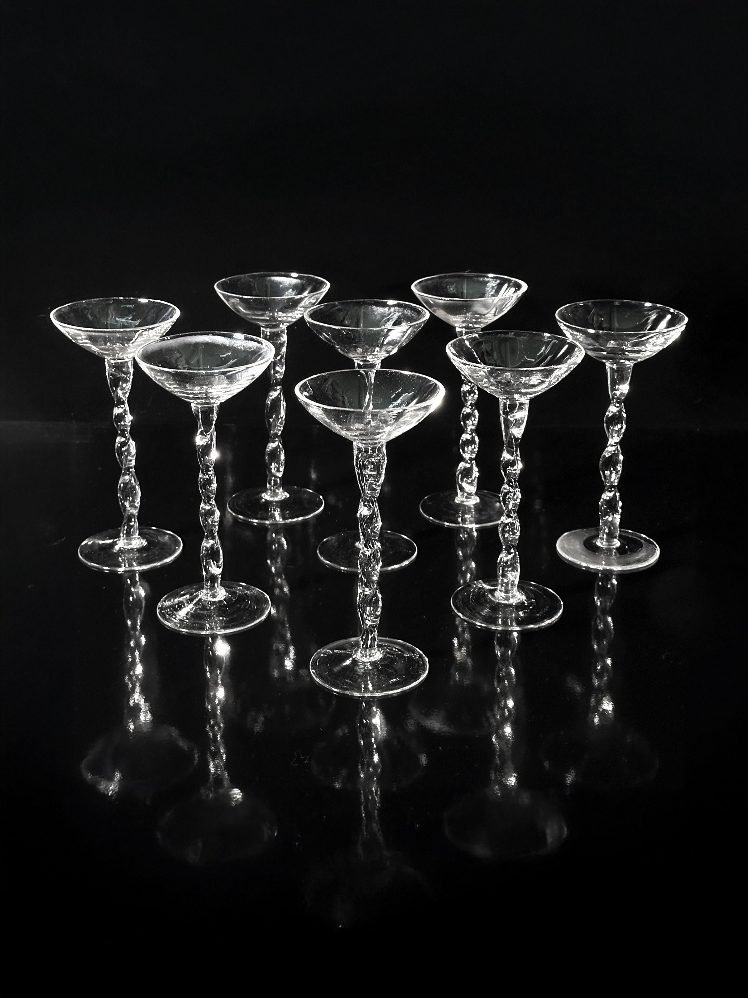 A set of 8 iridescent Dodo Vintage Art Nouveau liqueur glasses from the 1910s, featuring ornate twisted stems, are displayed against a dark background, casting reflective patterns below.