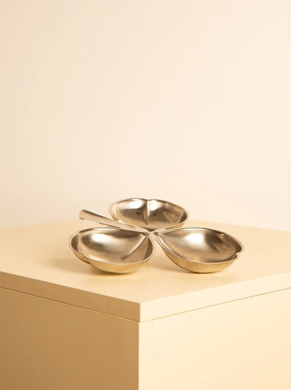 A metallic clover-shaped "Trefoil" shaped vide-poches 80's by Treaptyque with three connected sections and a handle, crafted from stainless steel. Placed on a light-colored wooden surface against a beige background, this '80s-inspired dish boasts a reflective, shiny finish.