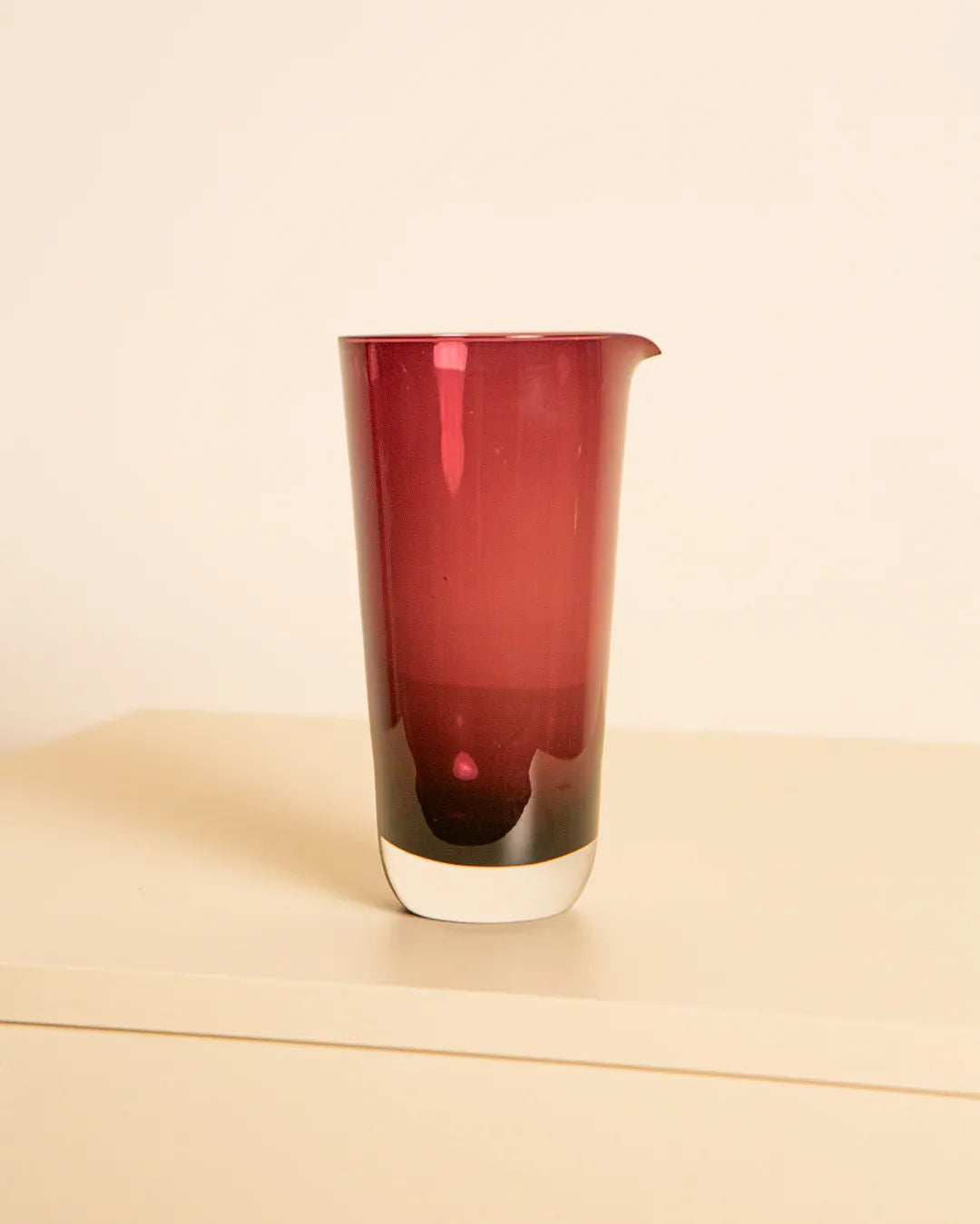 The Bordeaux Murano Glass Pitcher 80's by Treaptyque, a tall and elegant piece with a smooth surface and subtle spout, stands against a plain background. Its rich red hue showcases Italian artisanal glassmaking excellence, adding sophistication to the simple setting.