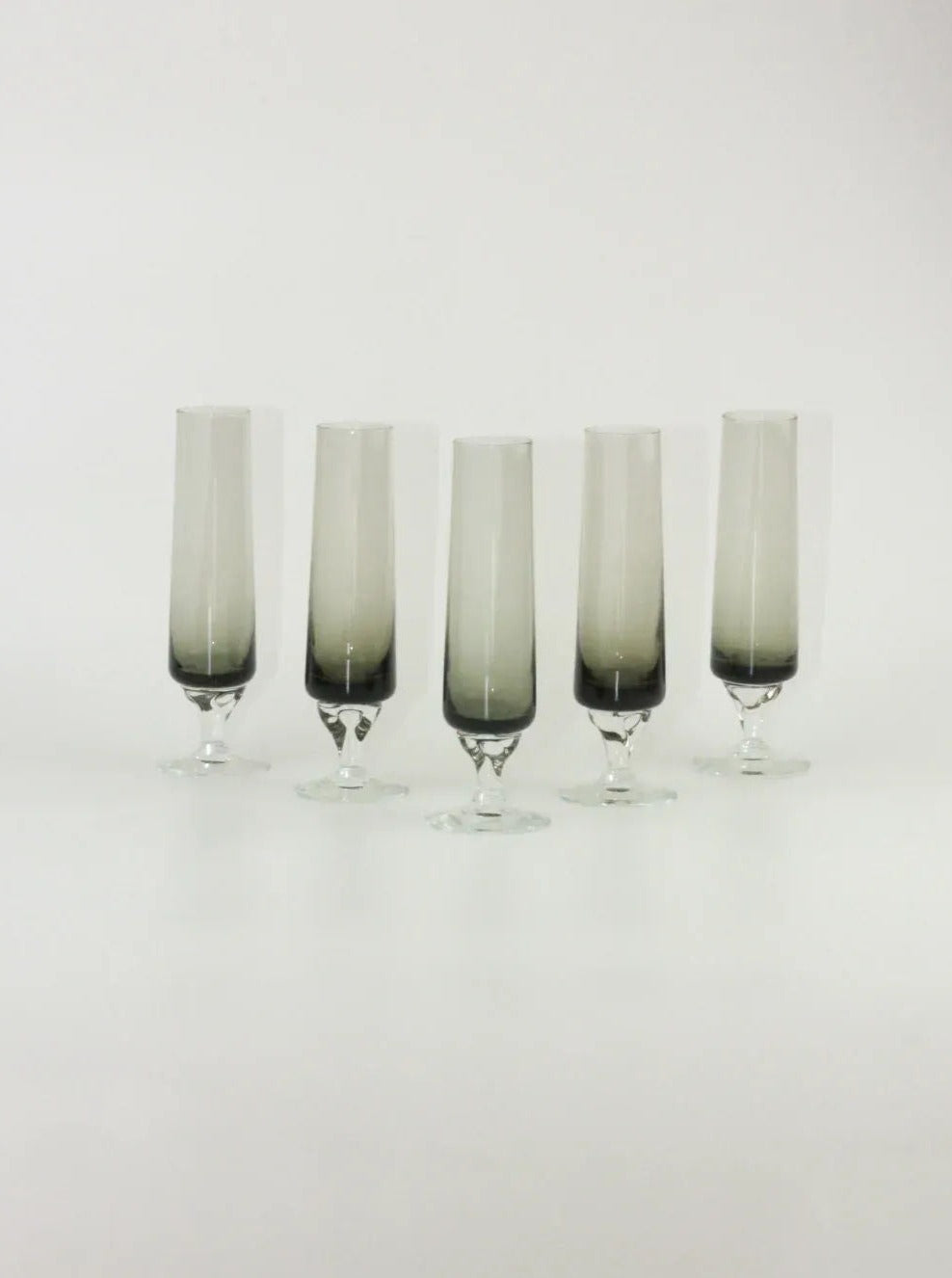 Five champagne flutes with gradient black-to-clear glass design and smoky gray stems, arranged in a row against a plain white background. The distinct tapering shape and elegant base highlight their modern aesthetic. This is the Set of Five Vintage Flute by Boga Avante Shop.