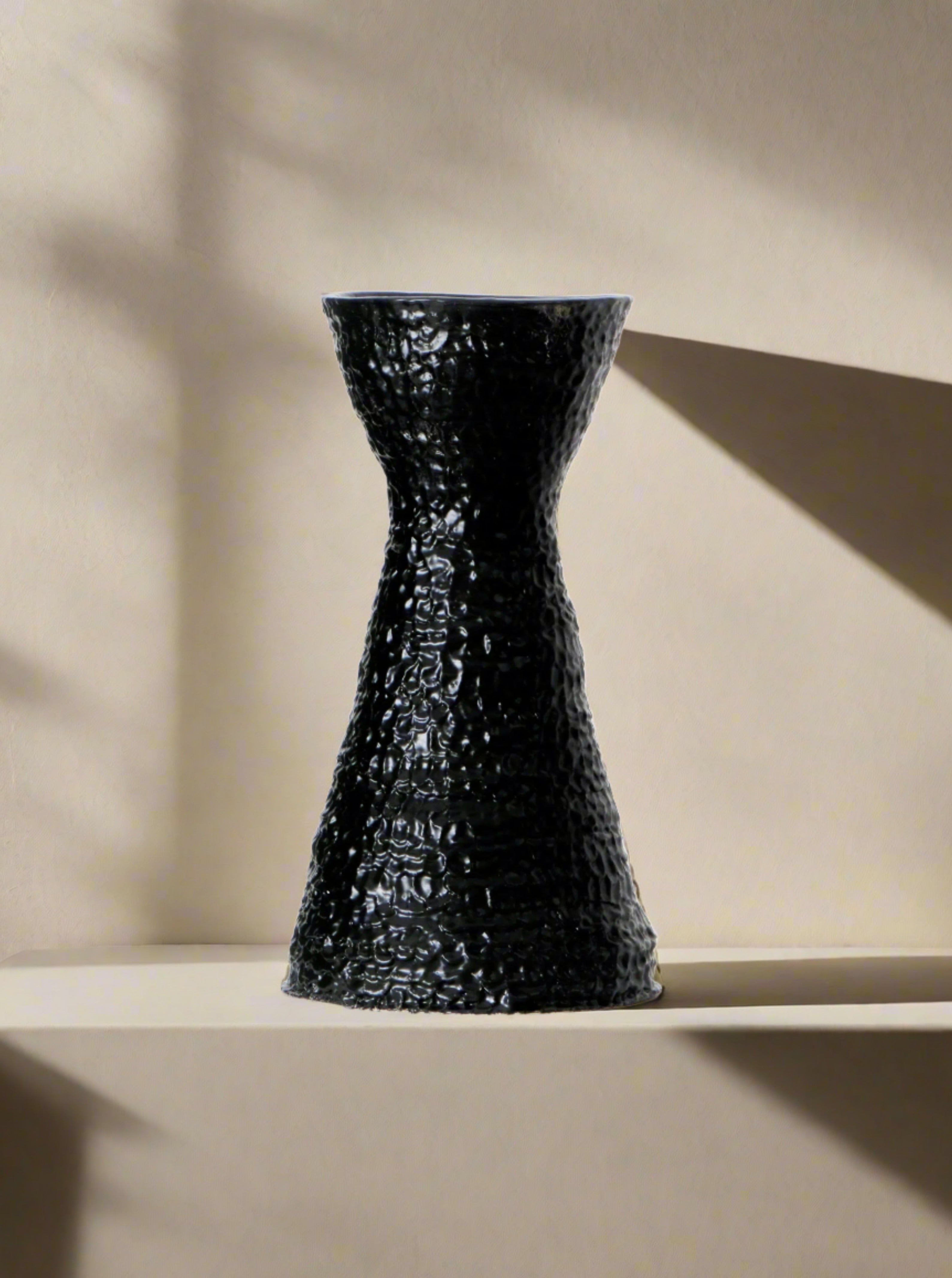 A black, hand-sculpted ceramic vase with a narrow middle sits on a white pedestal against a plain white background. The Casa Vase No1 by Project 213A features a unique, textured petal finish and an elegant hourglass shape.