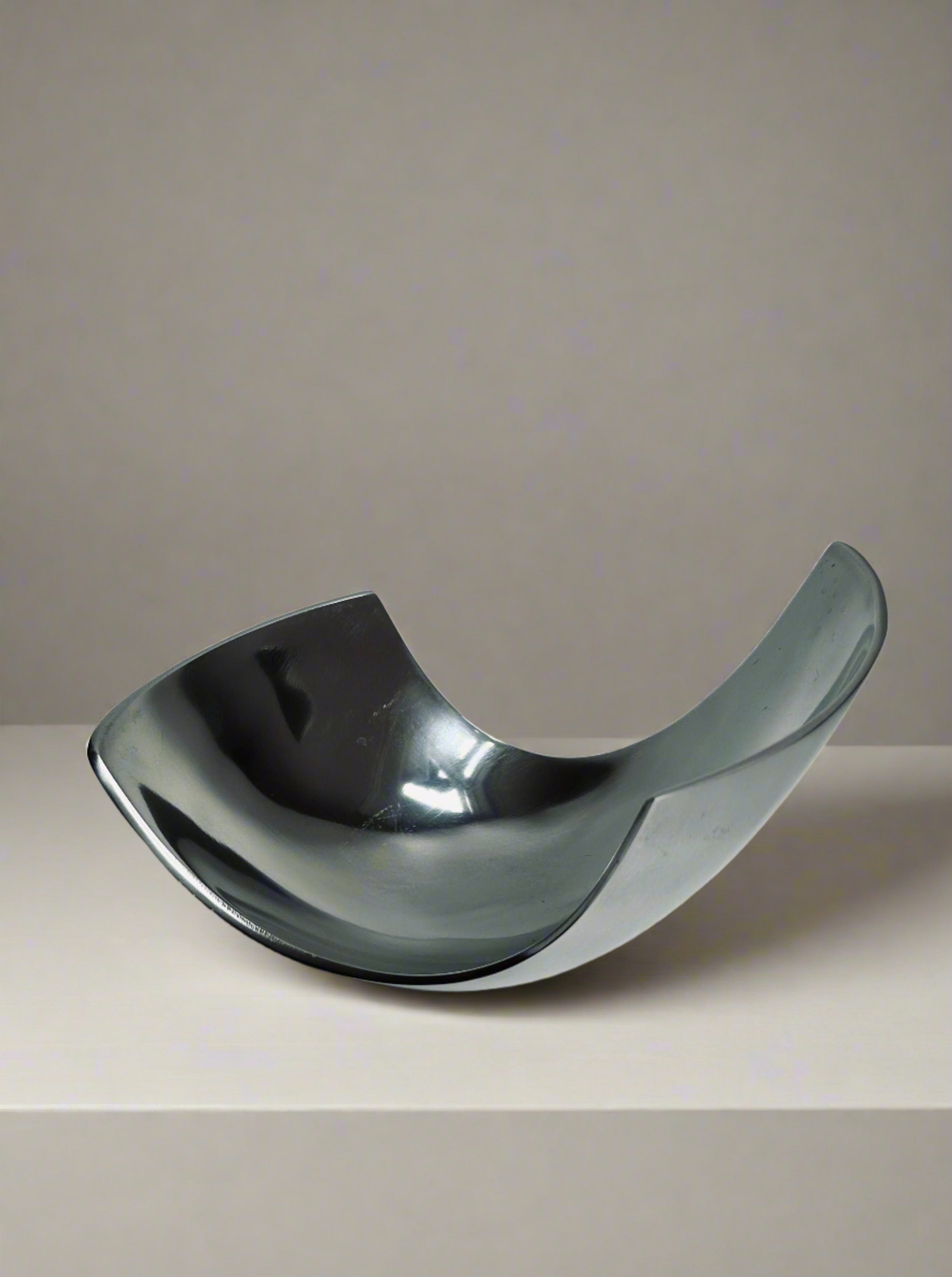 The Modernist Decorative Dish by Flock Studio features a sleek, reflective aluminum design with a distinctive curved shape, elegantly displayed on a plain surface against a neutral background. Its upward-curving sides create a modern, abstract look that is perfect for showcasing fruit.