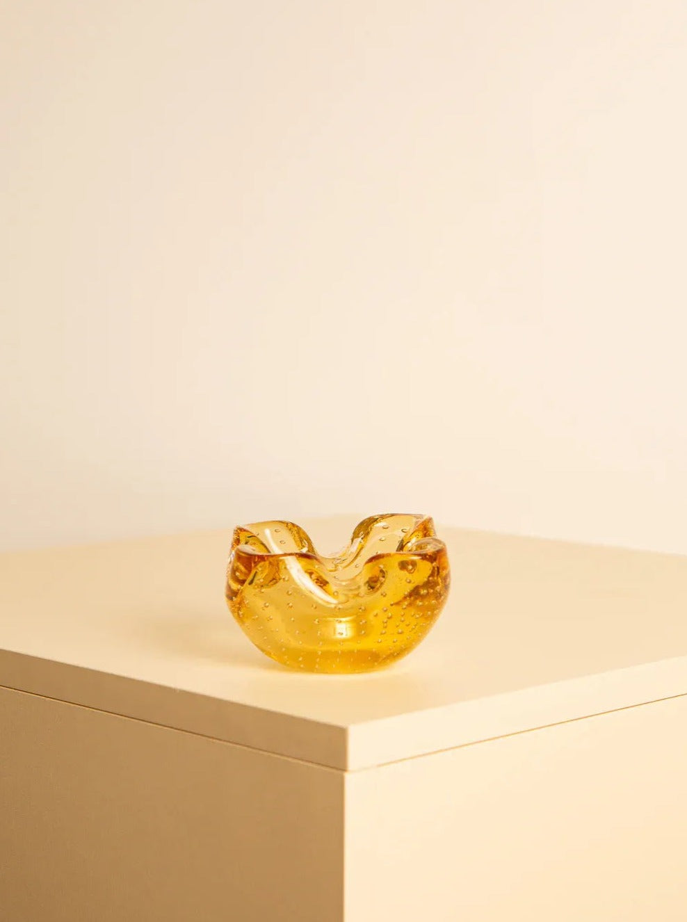 A small amber-colored glass bowl with a wavy, irregular rim sits on a light beige surface against a matching pale background. The bowl's translucent Murano glass catches the light, highlighting its smooth, glossy texture. The Vide-poches Murano 60's Yellow by Treaptyque adds a charming touch to any decor with its retro design.