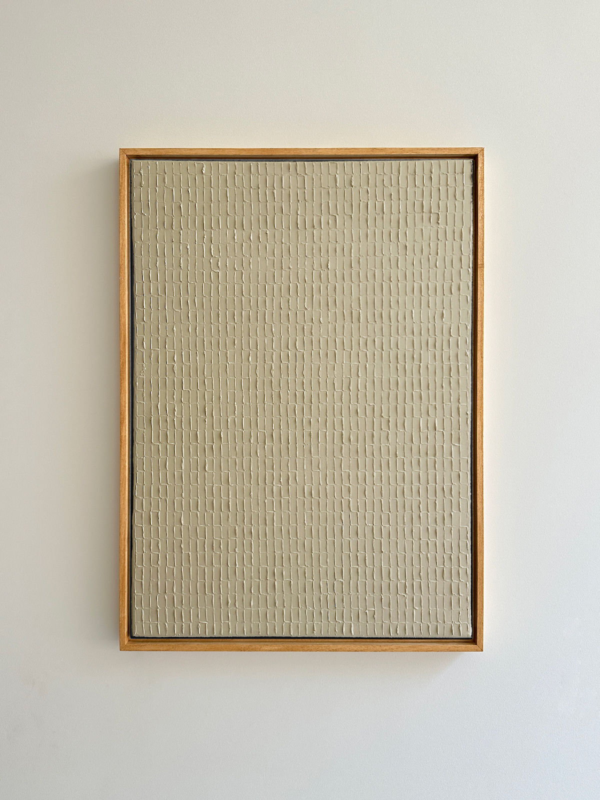 A minimalist artwork featuring a beige rectangular canvas with a textured, grid-like pattern, reminiscent of Korean Dansaekhwa. Buff Titanium - Oil on canvas by Roi Elmaliah, framed in a simple light wooden frame, exudes mindfulness and is displayed against a plain white wall.