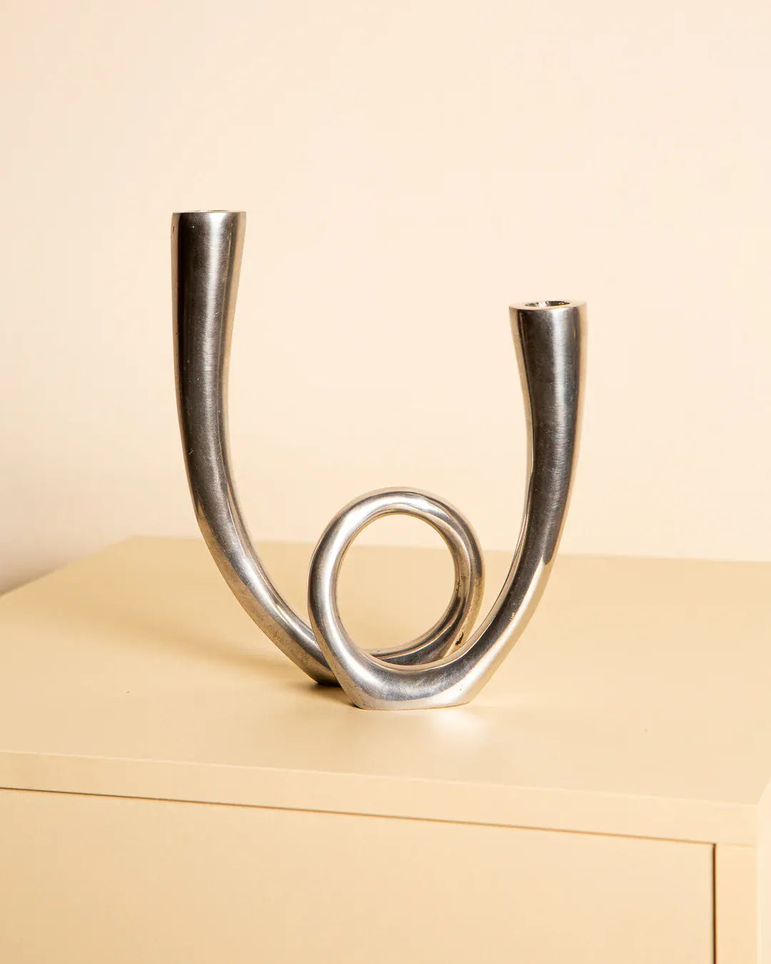 The Treaptyque Italian Candleholder in Cast Aluminum 80's showcases minimalist elegance with two tall, curved arms and a central circular loop. It rests on light wood against a beige background, blending modern abstract design with Italian artistry.