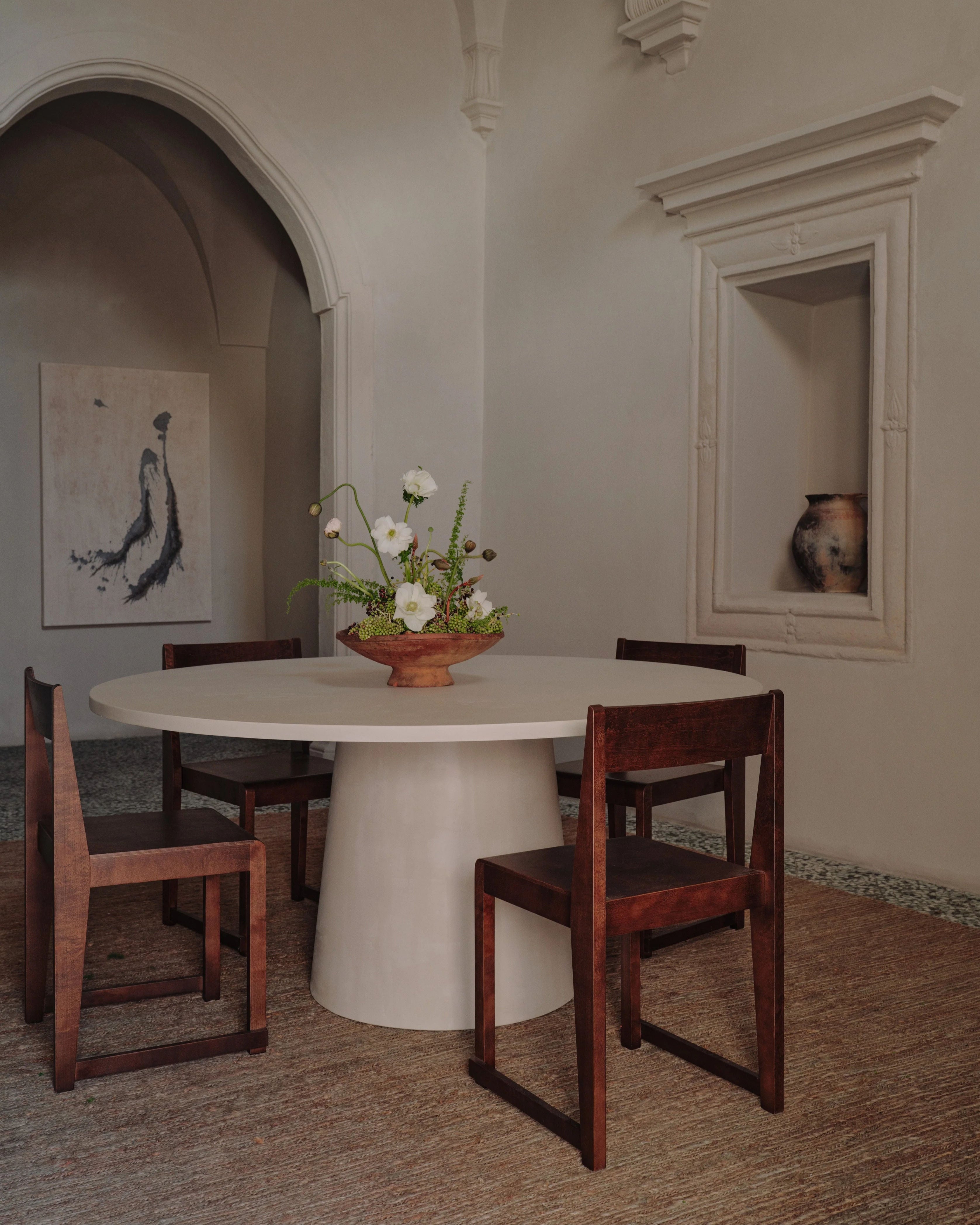 A minimalist dining room with a modern design features the MarlotBaus "Core" Dining Table, known for its scratch-resistant microcement top, surrounded by four wooden chairs and accented with a simple floral centerpiece. The room includes an arched doorway, a painting on the wall, and a niche showcasing a ceramic vase.