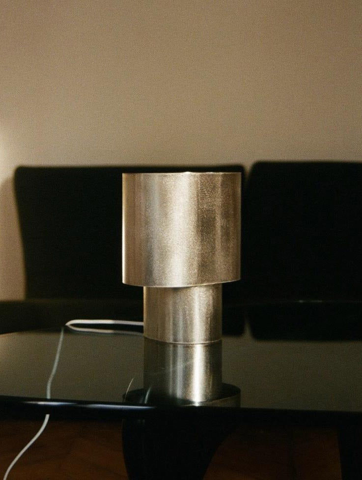 The Bien Paris Table Lamp in Brushed Steel, featuring a cylindrical design and white cord, sits on a glossy black table. A dark, blurred sofa and beige wall serve as the background.