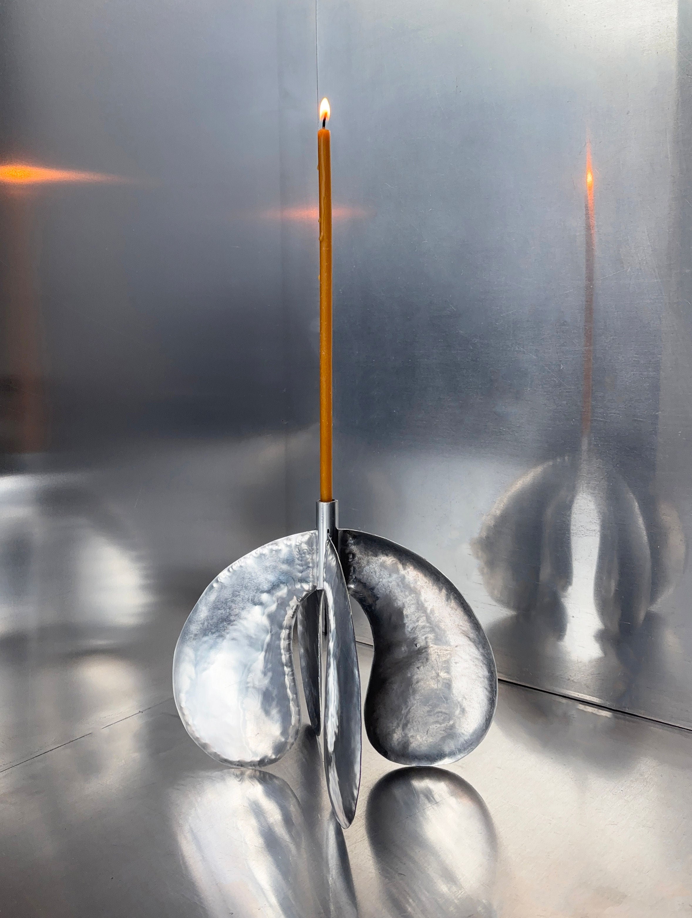 A tall, thin candle stands elegantly in the Phaedra Candle Holder Large by Bregje Sliepenbeek, handcrafted in Amsterdam with abstract curves. Its aluminum sheet base reflects its minimalist charm like a modern mirror.