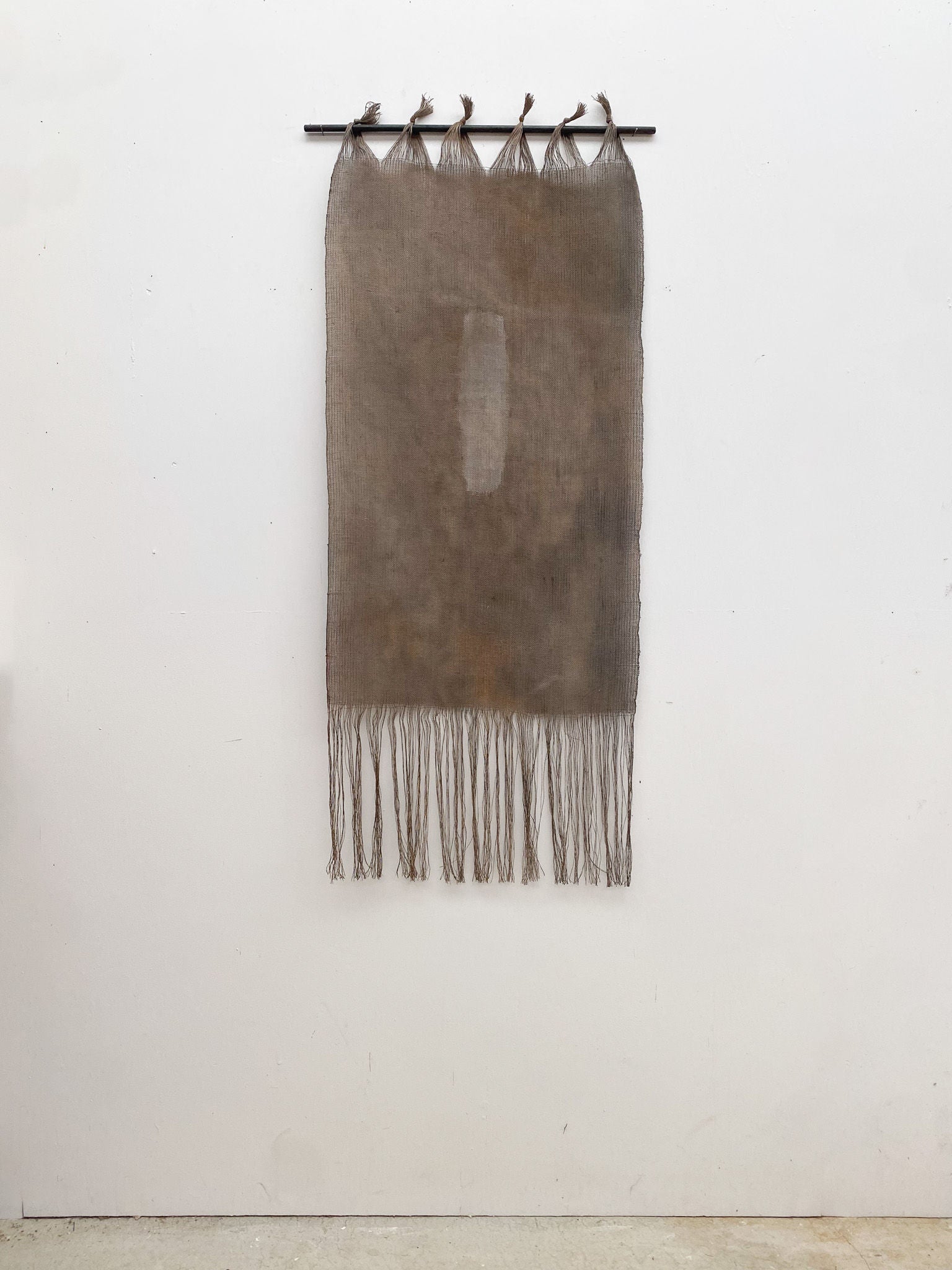 The Moontide tapestry by Sundance Studio hangs vertically from a black rod, showcasing Yuraygir National Park's beauty. Featuring fringes and a central rectangular patch secured by five loops, it stands out against the white wall like Yaegl Country's vast landscapes.