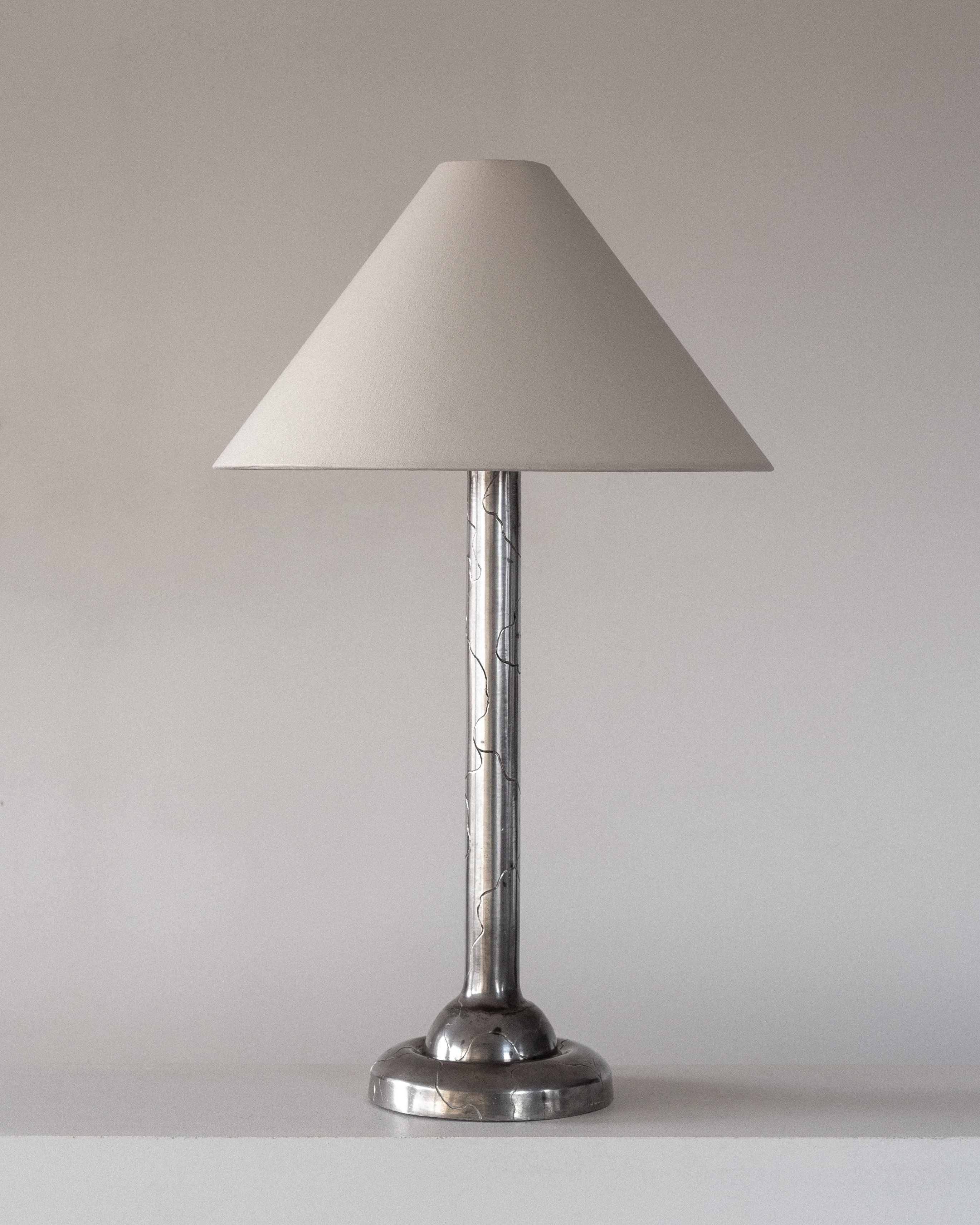 The Spigel Art Deco Pewter Lamp, inspired by 1920s Swedish design, features a pewter base and sleek stem. It comes with a conical beige shade that reflects 1920s elegance, set against a minimalist light background.