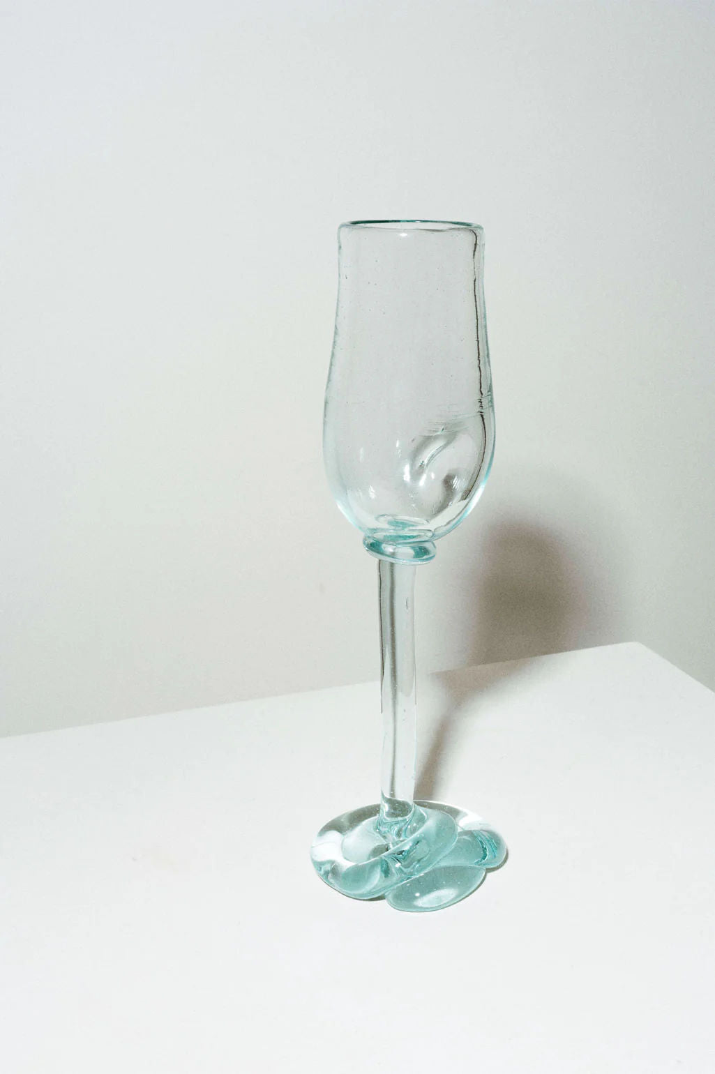 The Thaw - Recycled Flute Glass in Clear by Completedworks features a transparent, uniquely twisted design crafted from recycled glass. It stands on a white surface with an artistically shaped irregular base and a straight, elongated stem against a plain light gray wall.