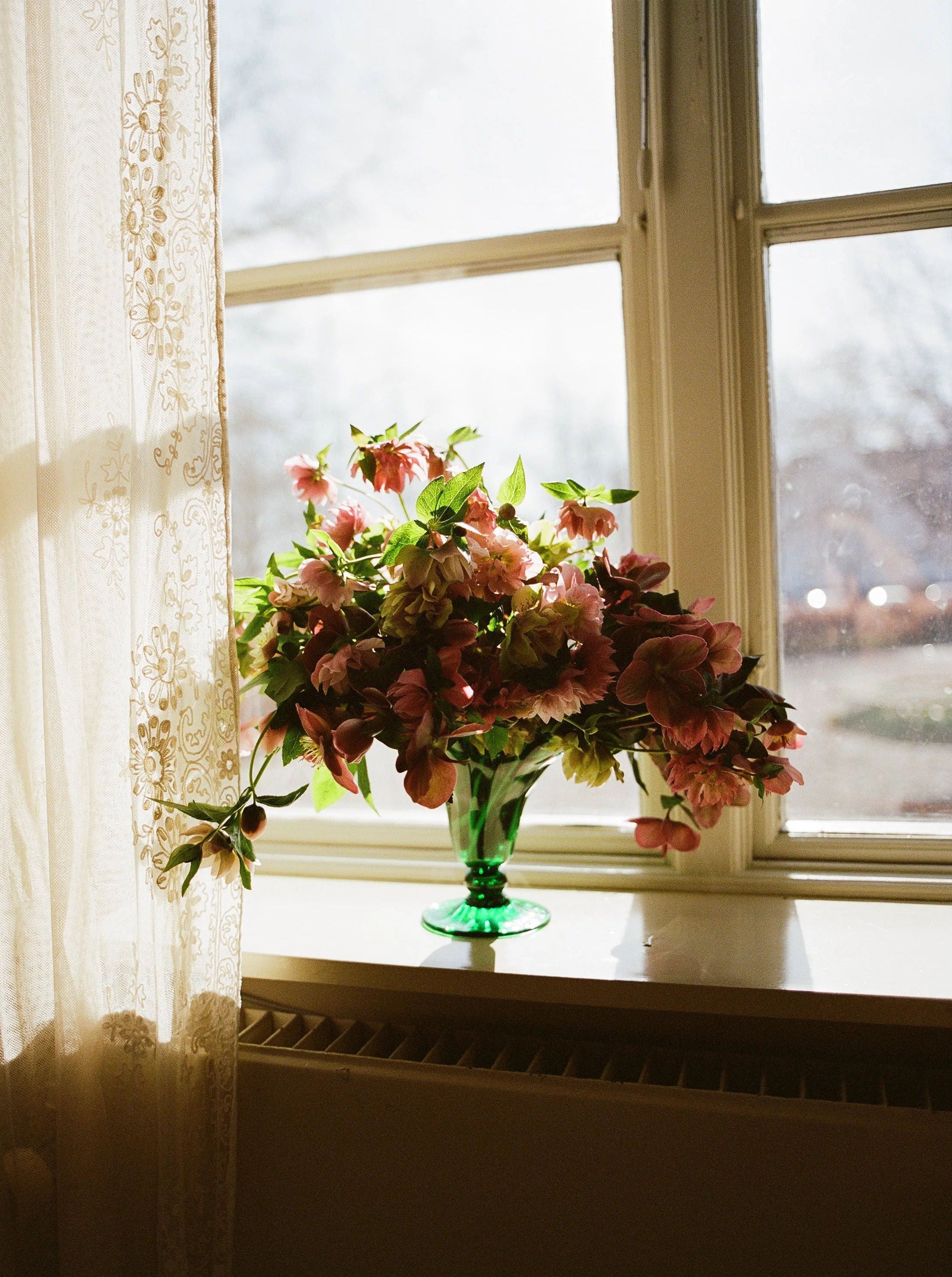 A green Karen Blixen Vase Small by Akua Objects filled with pink flowers is placed on a sunlit windowsill. The window is partially covered by a sheer lace curtain, and the view outside is slightly blurred, suggesting a bright, sunny day.