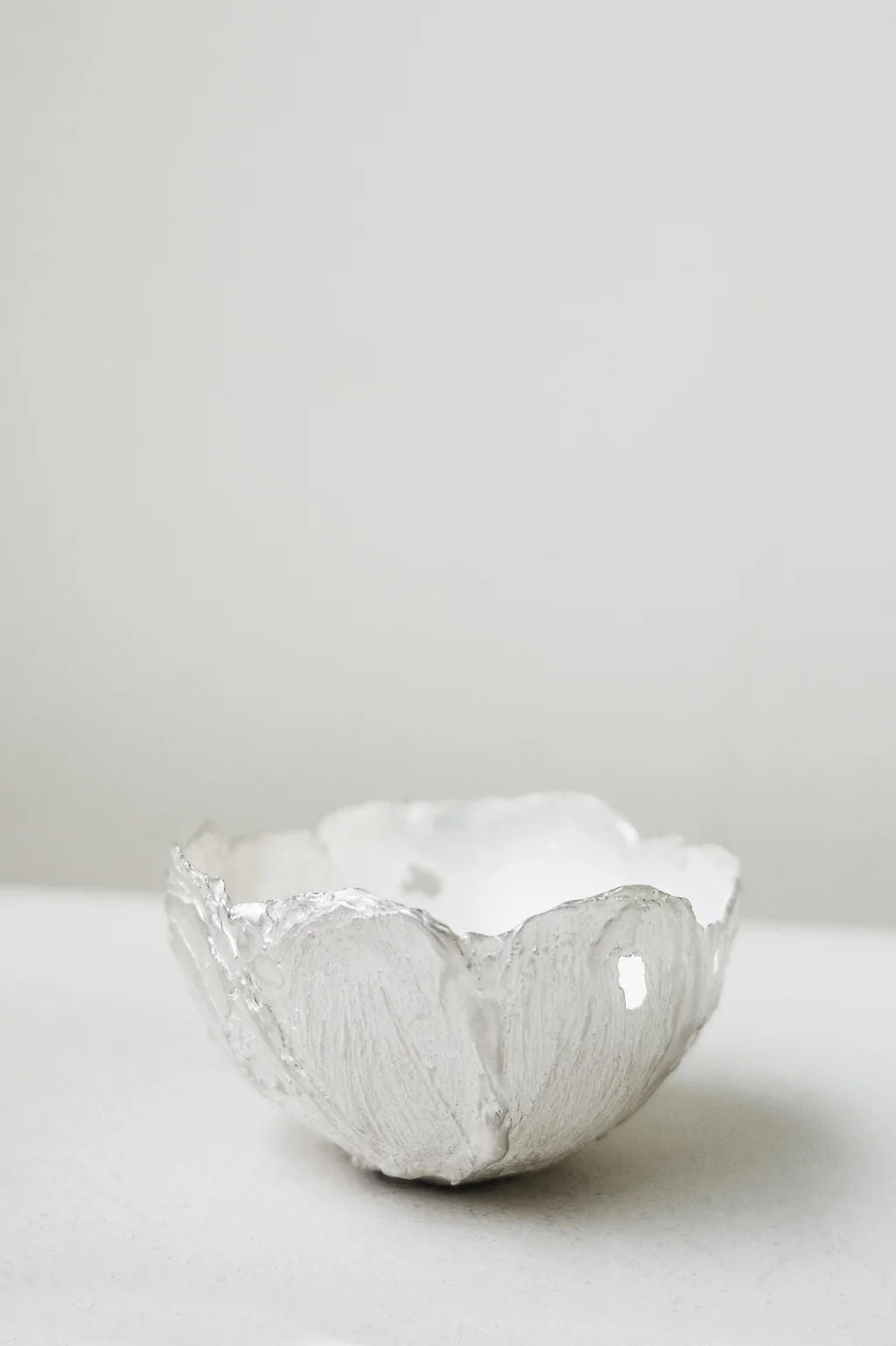 The L34 - Small Bowl in Silver by Completedworks, with its textured white ceramic and organic shape, rests on a smooth white surface against a plain light gray background, highlighting its charm as a UK handmade piece.