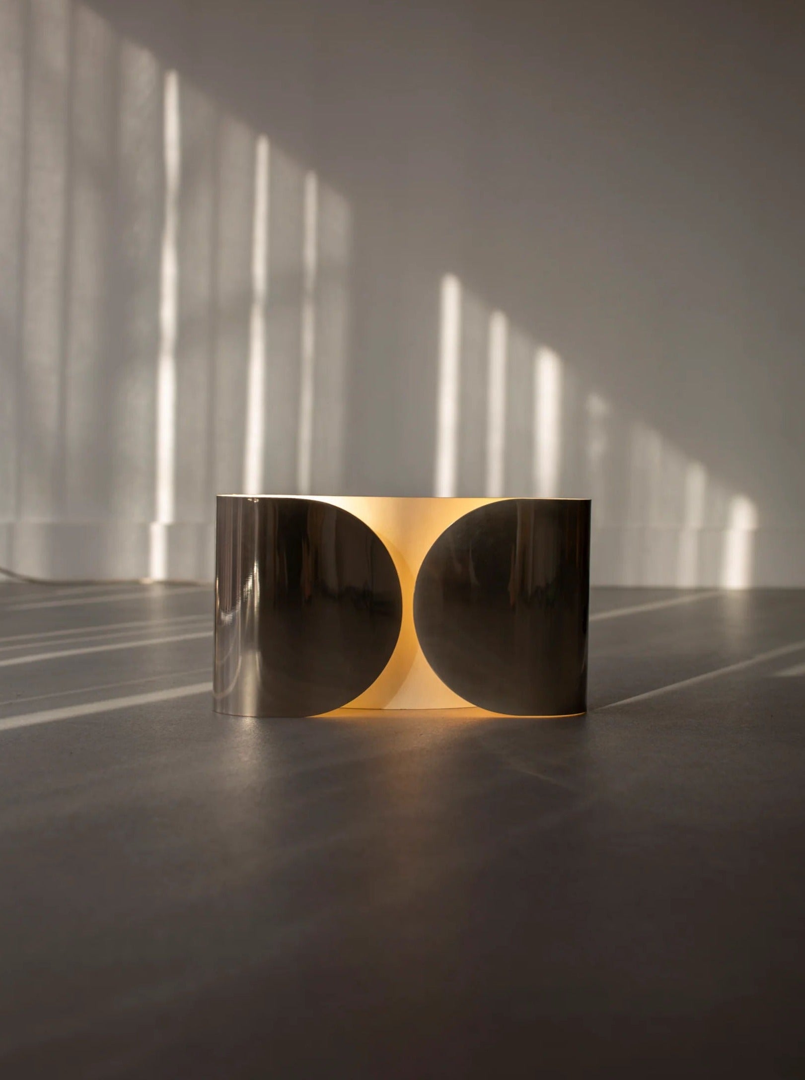 A modern, circular light fixture emits a warm glow, casting gentle shadows on the floor. Sunlight filters in through sheer curtains in the background, highlighting a sleek Chrome Wall Lamp by Tobia Scarpa 80's from CU-RA-TED and creating a serene and minimalist ambiance in the room.