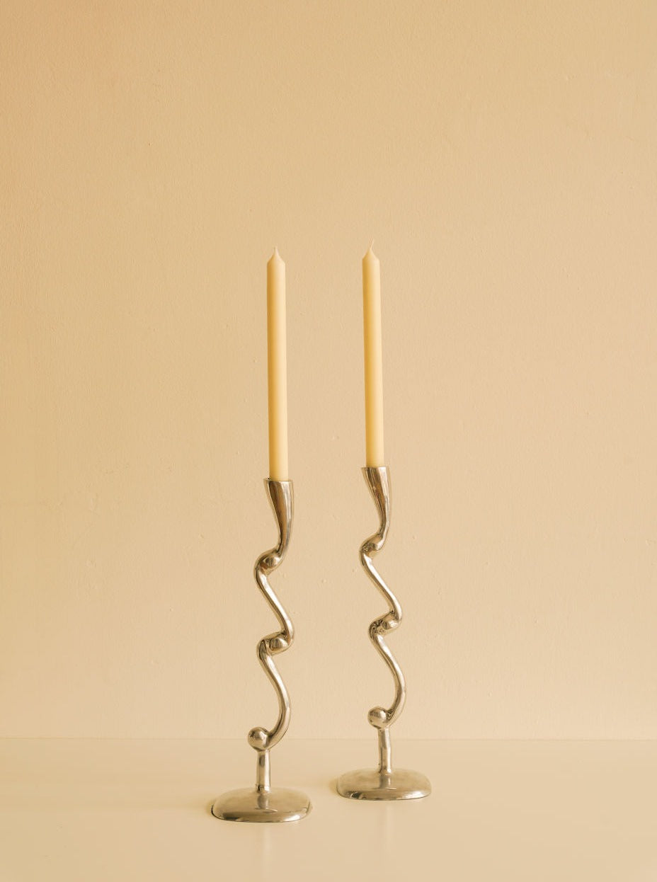 Two tall, slender candles in elegant, Les Objets Amsterdam Curly Candleholders stand against a plain, light beige background.