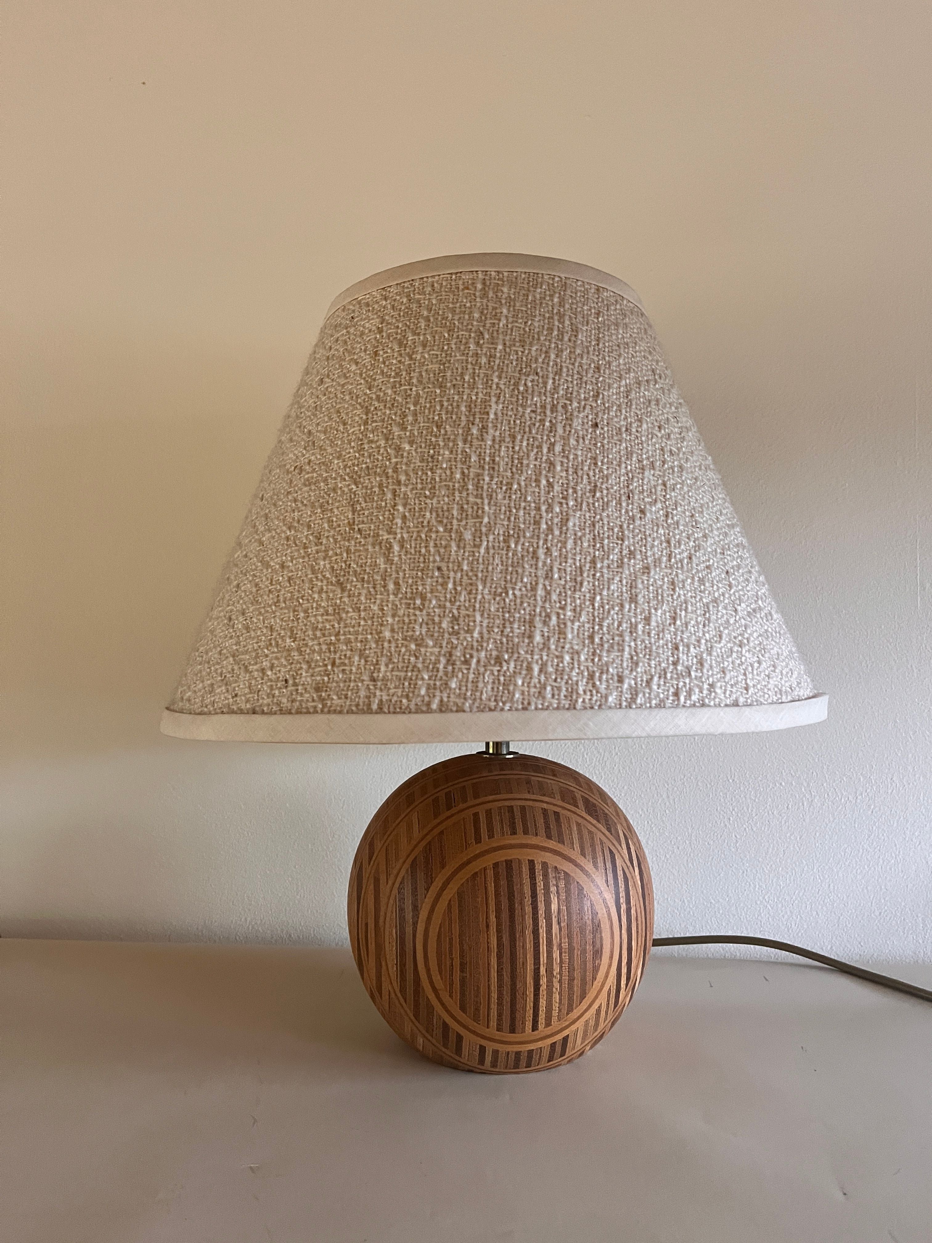 The Maud Vaughan Wood Table Lamp 1970s, featuring a vintage boucle shade and a wooden spherical base adorned with circular patterns, sits elegantly on a light-colored surface against a plain wall.