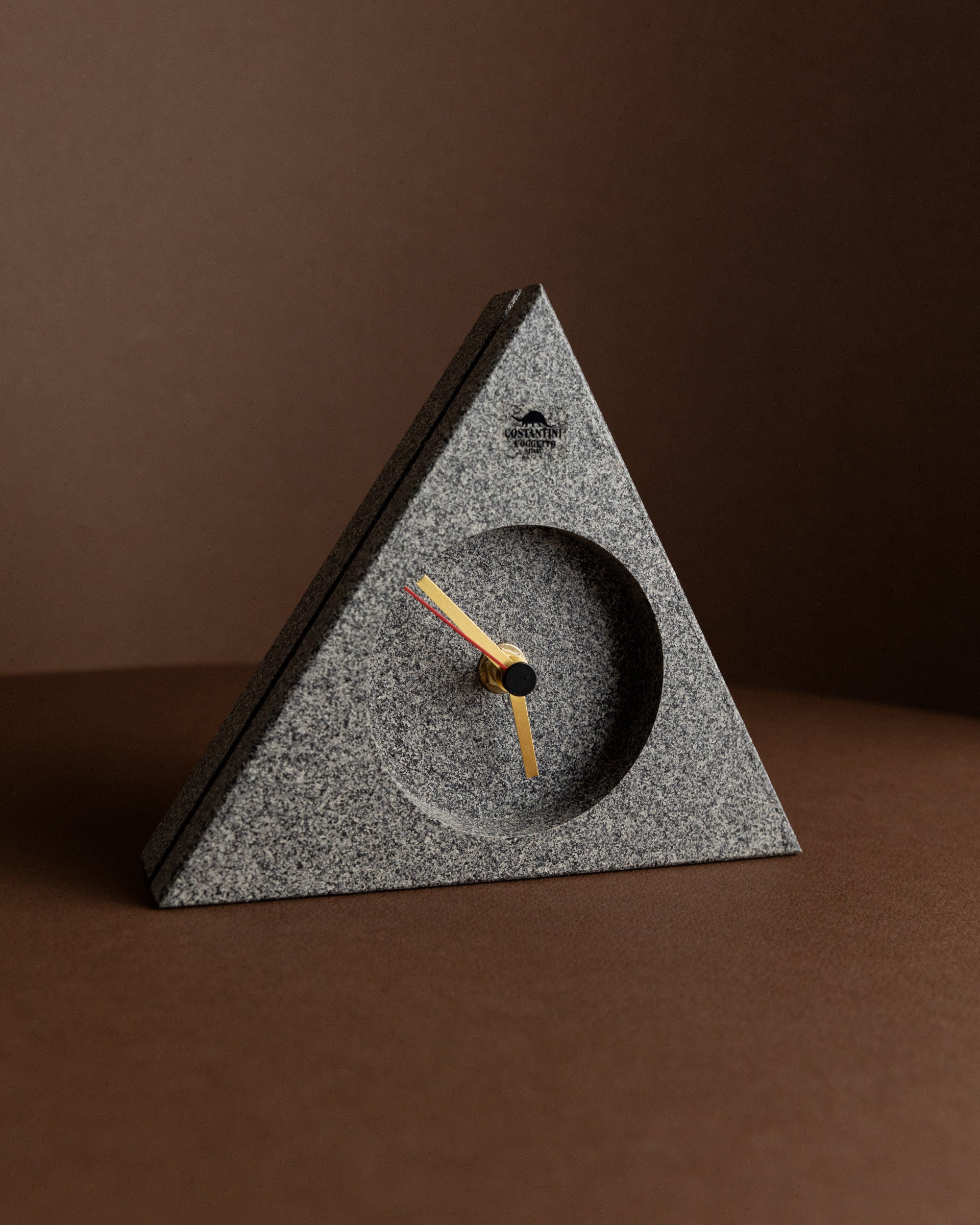 A Bottega Jacobs Vintage Costantini l'Oggetto grandfather clock, featuring a triangular design in speckled gray plastic, embodies a minimalist aesthetic. The clock includes black hour and minute hands, a contrasting orange second hand, and is set against a plain brown backdrop. Crafted in Italy with 1980s postmodern Memphis style pop art influences, it's an ideal retro gift idea.