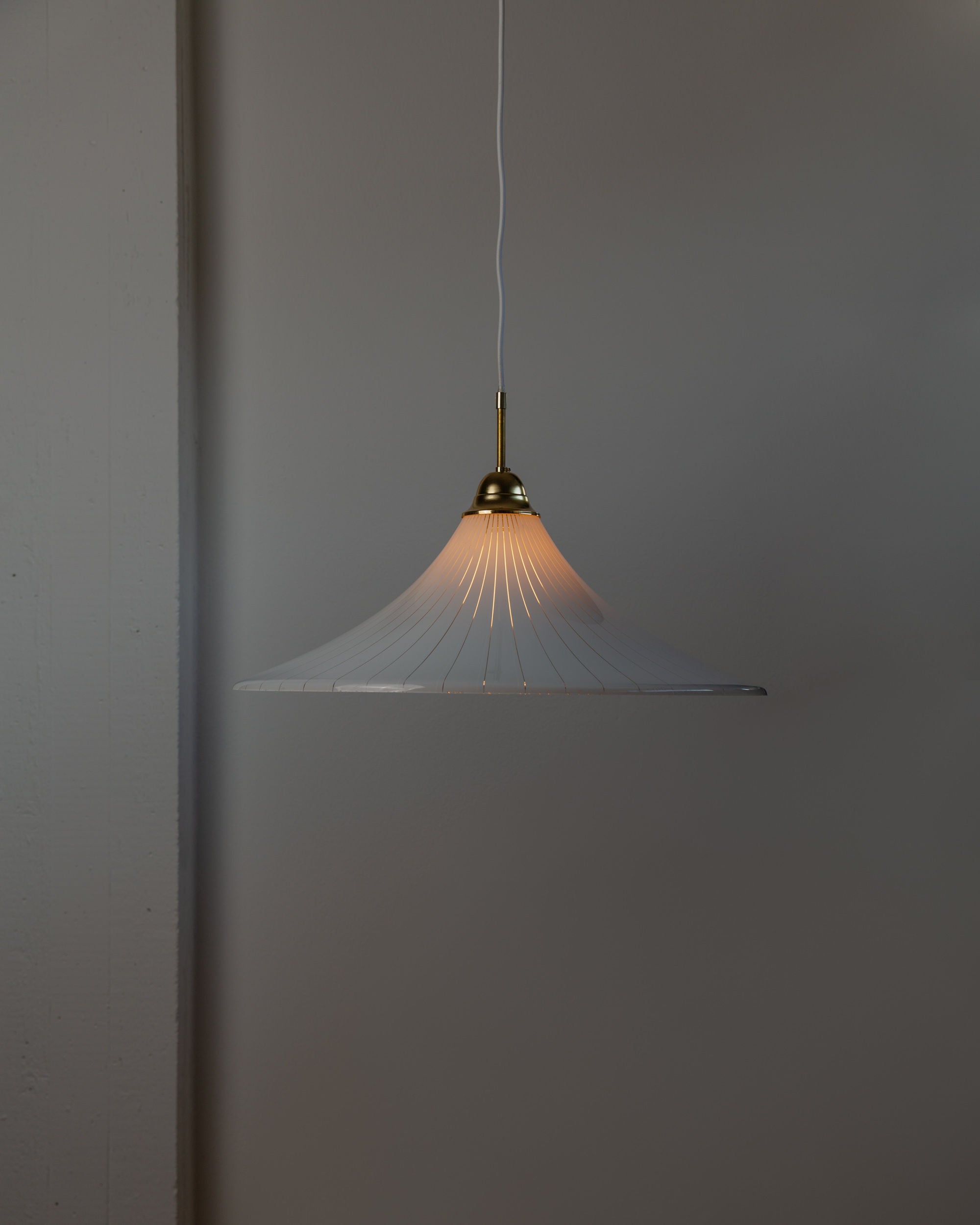 A minimalist Ceiling Lamp 1970s Italy from Bottega Jacobs, featuring a wide, flared white lampshade, hangs elegantly from the ceiling, creating an XL vintage ceiling lamp ambiance. The light is turned on, softly illuminating the plain dark gray wall in this modern interior setting.