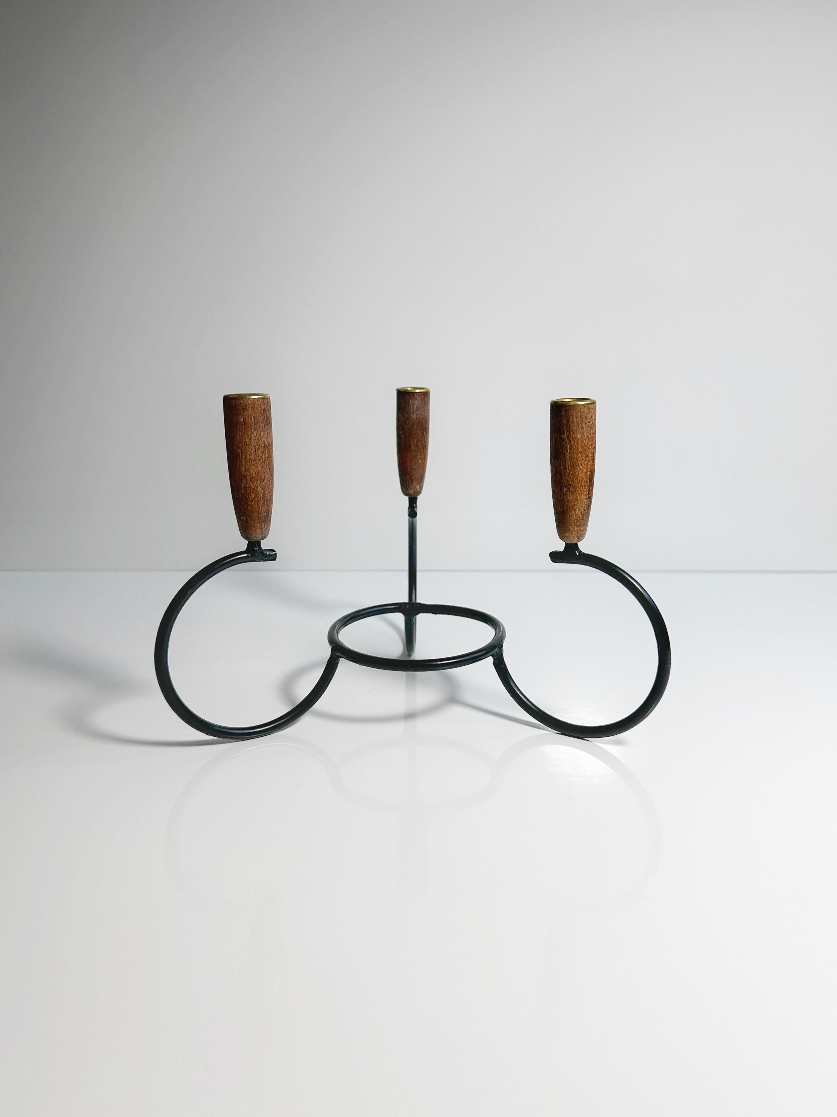 The Danish Candleholder 1960s by Dodo Vintage is a minimalist black metal candle holder that features three circular bases, each topped with teak ends. Designed to hold three candles, it is set against a plain, light background to create a clean and simple aesthetic.