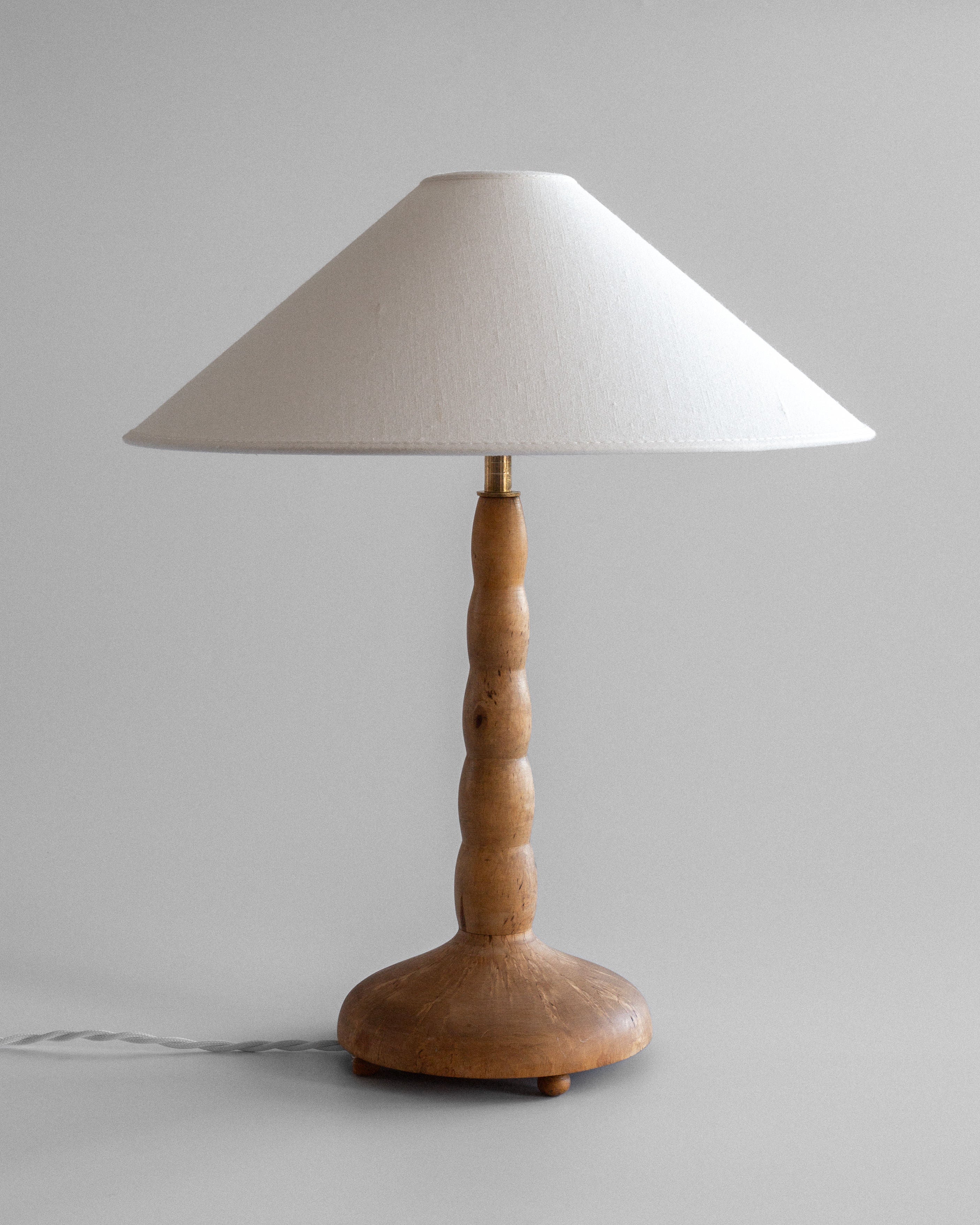 The Spigel Turned Birch Table Lamp from Sweden features a white conical lampshade and wooden base, showcasing exquisite craftsmanship. Its unplugged design with the cord visible against a plain gray background adds elegance to any room.