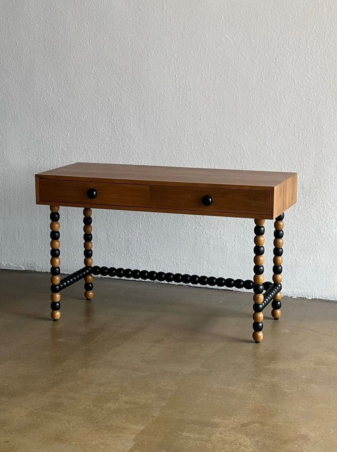 The Onde Table by Studio Kallang features a two-toned finish and two drawers with black knobs. The playful desk stands on four spiral legs composed of alternating black and wood-colored spheres. It is placed against a plain, white wall on a smooth, beige floor.