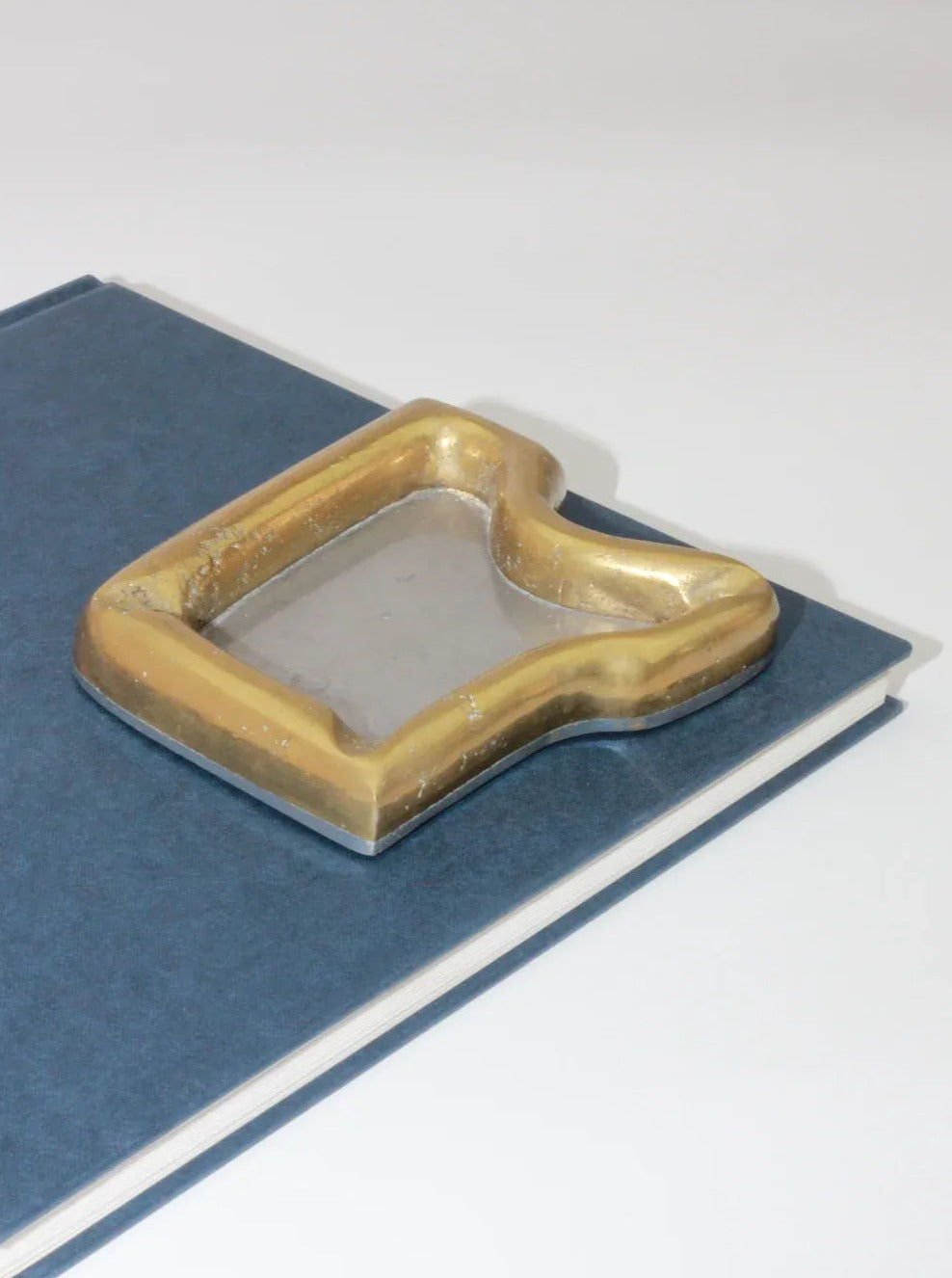 A book with a dark blue cover sits on a white surface. Atop the book rests the Brutalist Ashtray by Boga Avante Shop, with its quirky, rectangular shape featuring a recessed area and worn edges. This vintage item adds an intriguing touch to the scene.