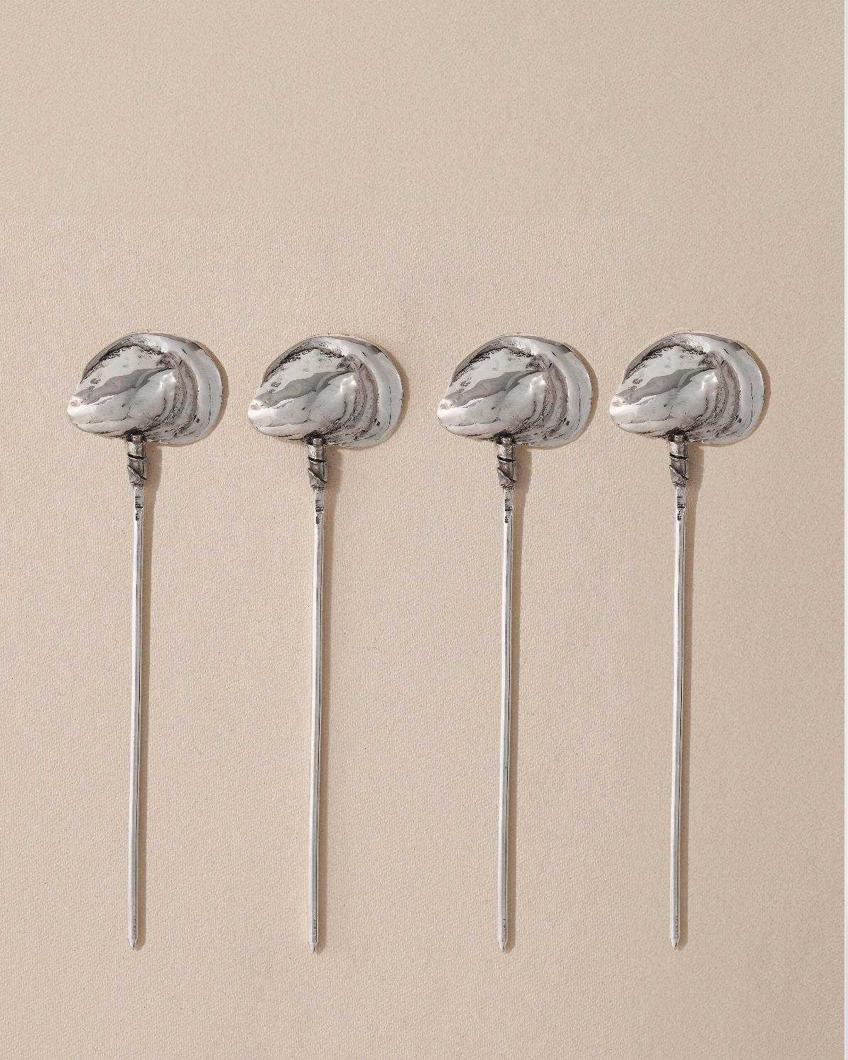 A set of four Oyster Cocktail Picks from Heath Wagoner - Artisanal Cutlery and Objet D’art is elegantly laid out on a beige surface, each featuring a meticulously detailed oyster shell design that radiates maritime sophistication.
