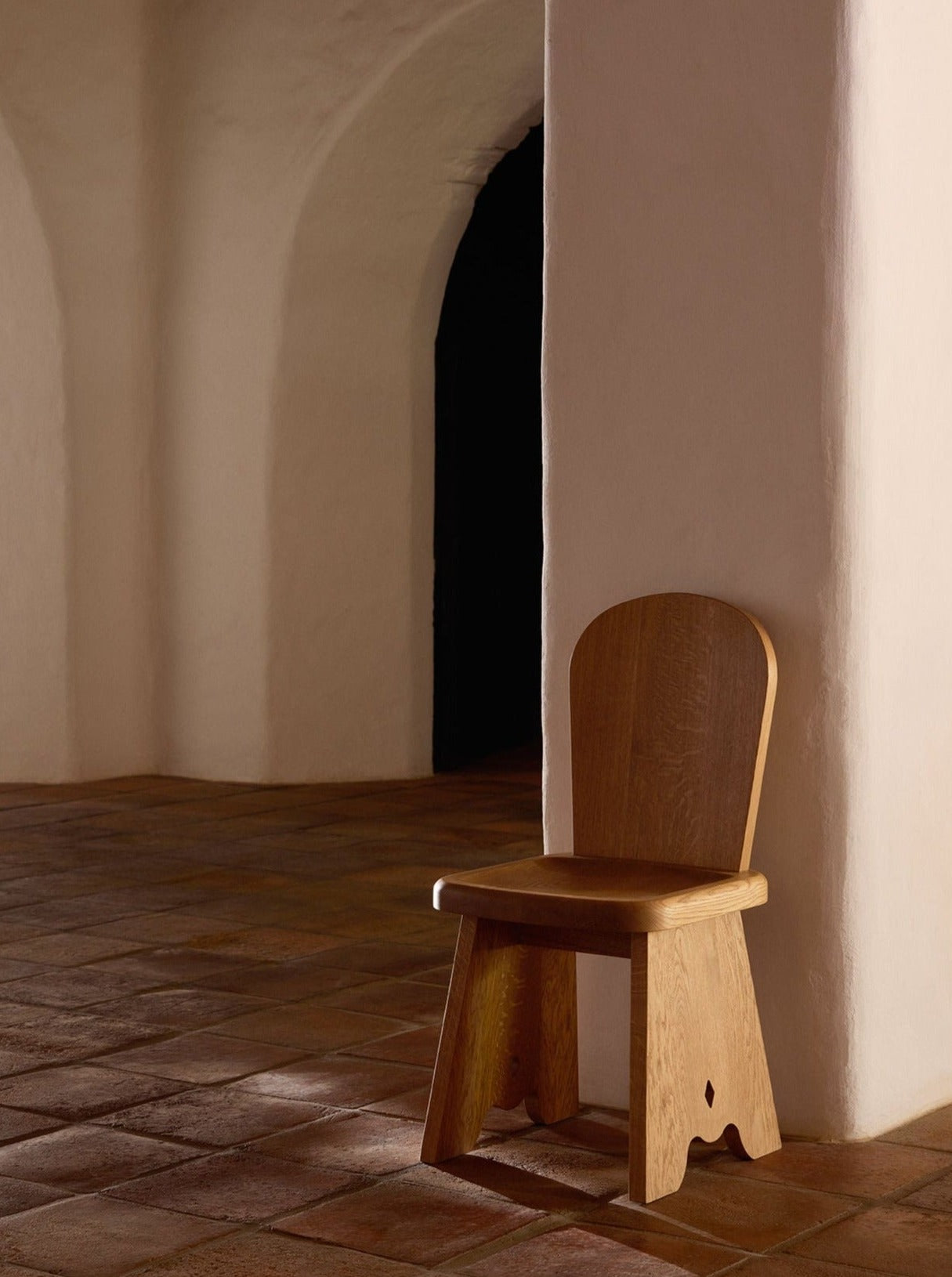A Lemon Rambling Chair in Natural French Oak Wood, with a simple, rounded backrest is placed against a white, curved wall in a room with terracotta floor tiles. The space features arched doorways, adding an element of architectural interest to the serene, minimalist setting.