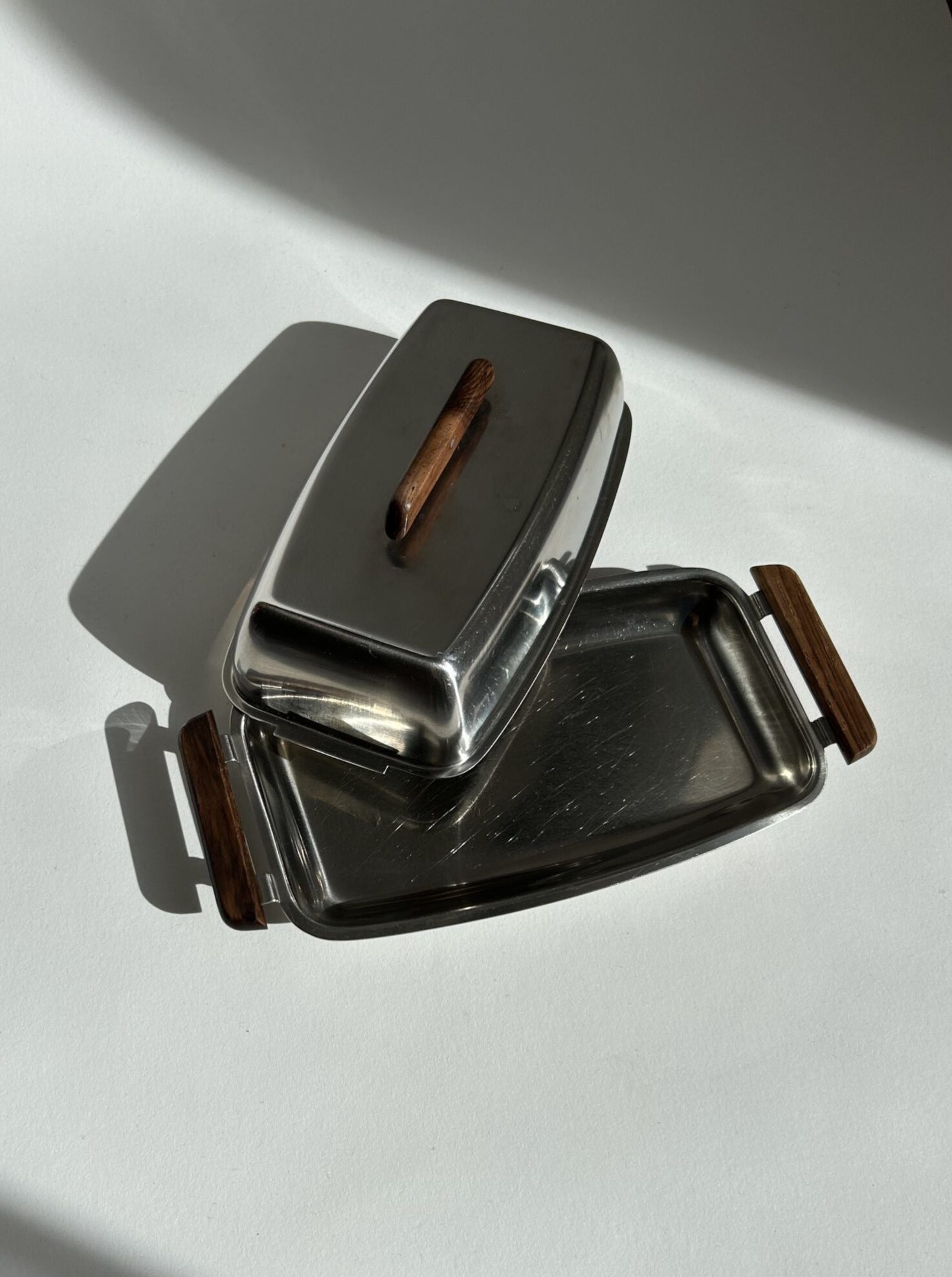 The Mid Century Butter Dish by Stelton from Flock Studio, featuring a stainless steel body and a rosewood handle, encapsulates the essence of mid-century design allure. When positioned slightly askew on its base, it projects a gentle shadow onto the white surface, offering a minimalist yet vintage modern appeal.