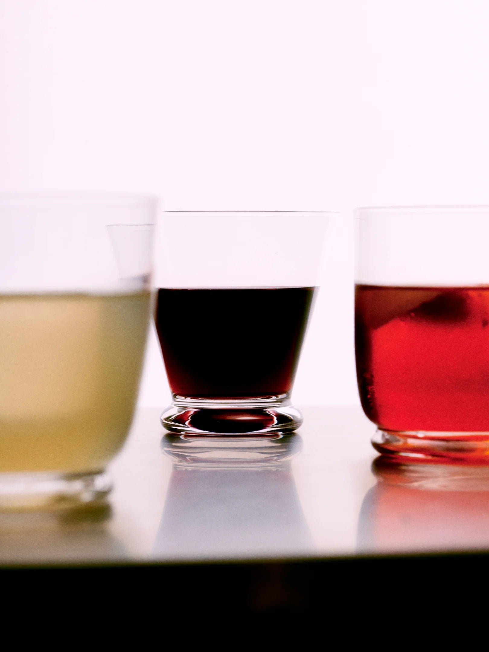Three tumblers from the "Walter 03 - Coupé / Set of 4" by Service Projects, meticulously crafted from lead-free crystal and mouth-blown to perfection, are filled with different colored liquids. A light yellow-green beverage sits on the left, a dark red drink in the center, and a reddish drink with ice cubes on the right—all placed on a reflective surface against a plain white background.