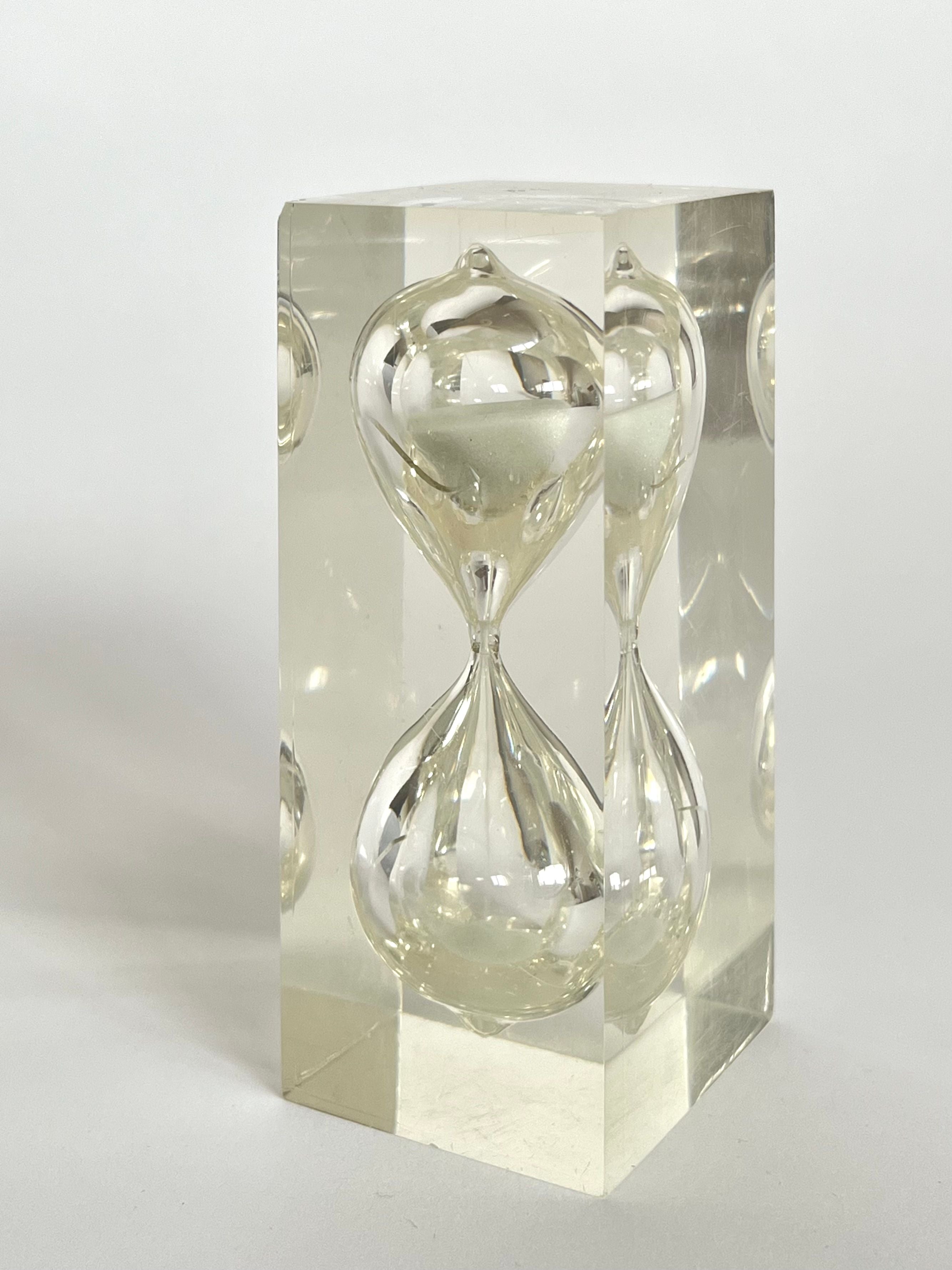 A clear, rectangular lucite cube encases the "Hourglass by Charles Hollis Jones, 1970s" from septembre studios, creating an illusion of multiple reflections. The hourglass appears suspended at the center, framed by smooth, transparent surfaces reminiscent of Jones’s innovative designs.