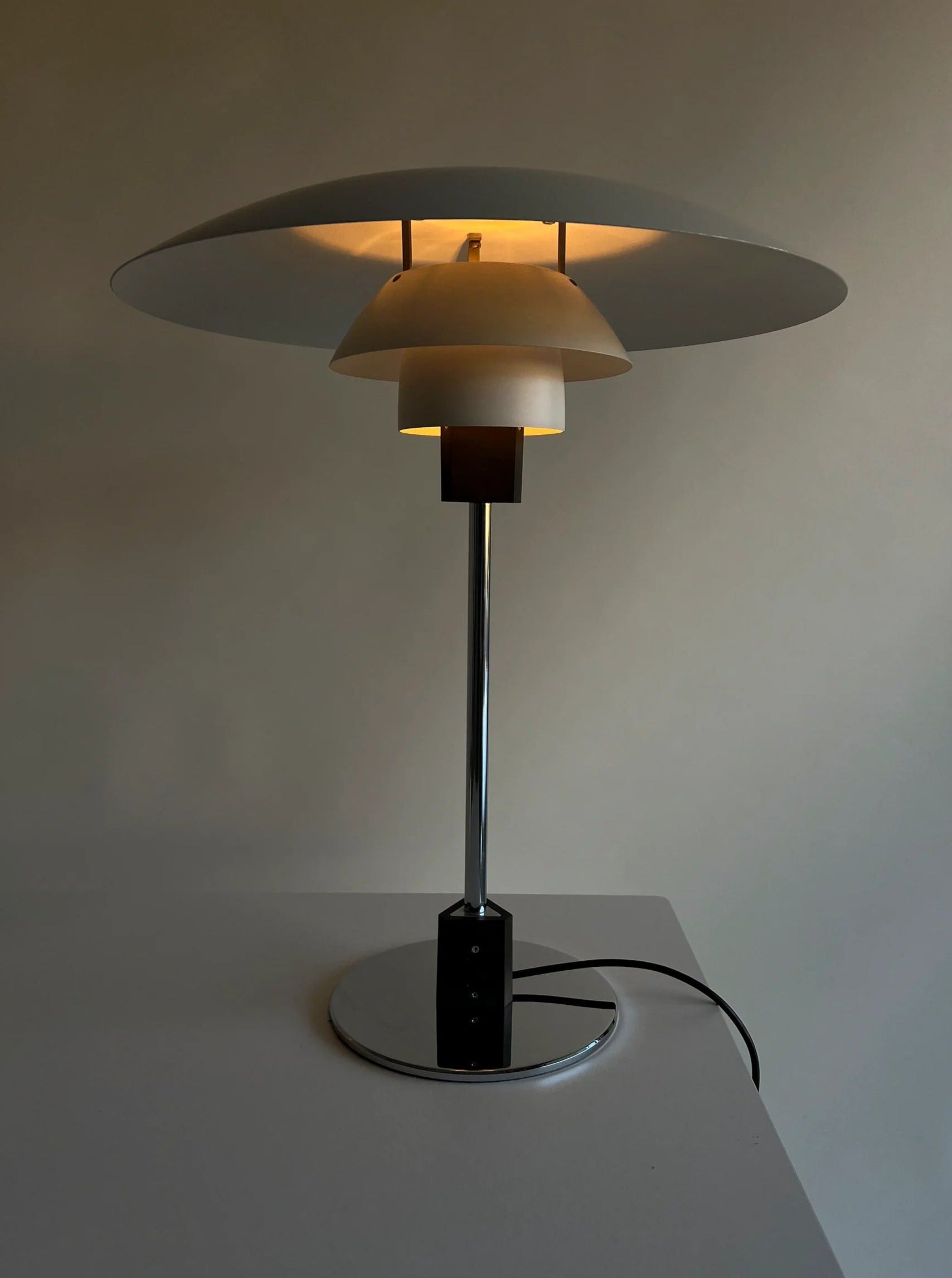 A modern CU-RA-TED Poulsen Table Lamp PH 4/3 1983 with a minimalist design reminiscent of Poul Henningsen’s work. It features a polished chrome stand and a multi-layered, outward-curving white shade that emits a soft, warm light. The lamp is situated on a sleek, white surface with a visible power cord.