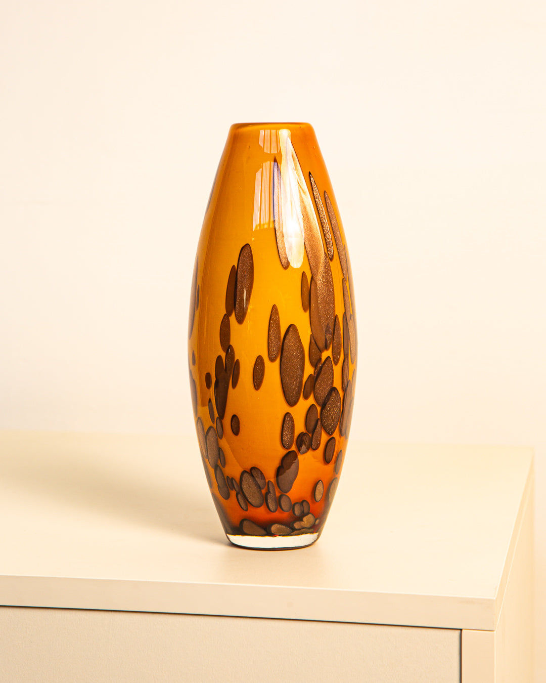 The Treaptyque Murano Blown Glass Vase 80s, a tall, oval orange vase with brown irregular spots, rests on a light surface against a plain, light background.