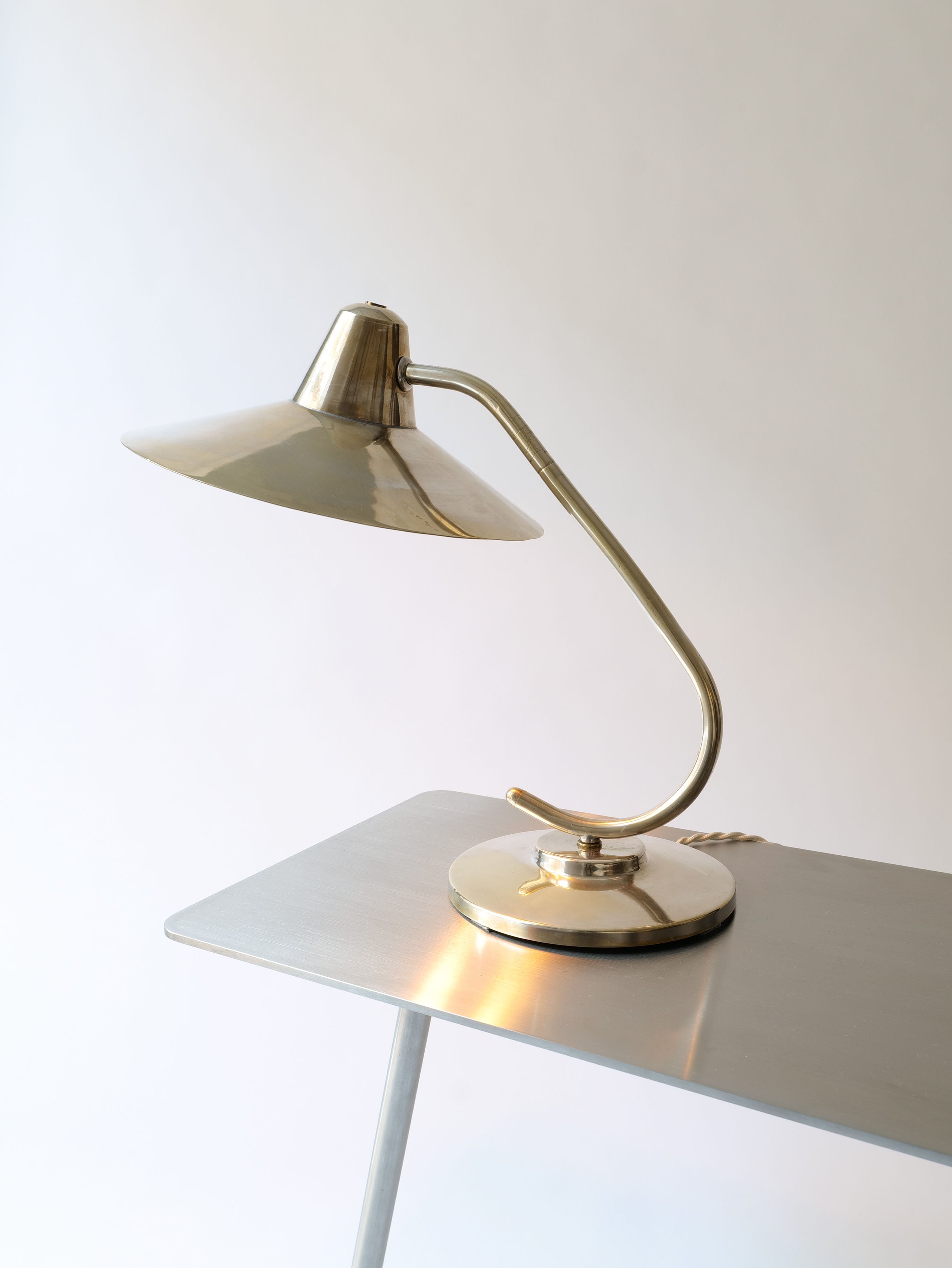 A sleek, midcentury Silver Brass Desk Lamp with a silver-plated brass finish by Collection Apart sits on a minimalist white desk. The lamp features a curved neck and a wide, conical shade directing the light downward. The scene is set against a plain, light-colored background.