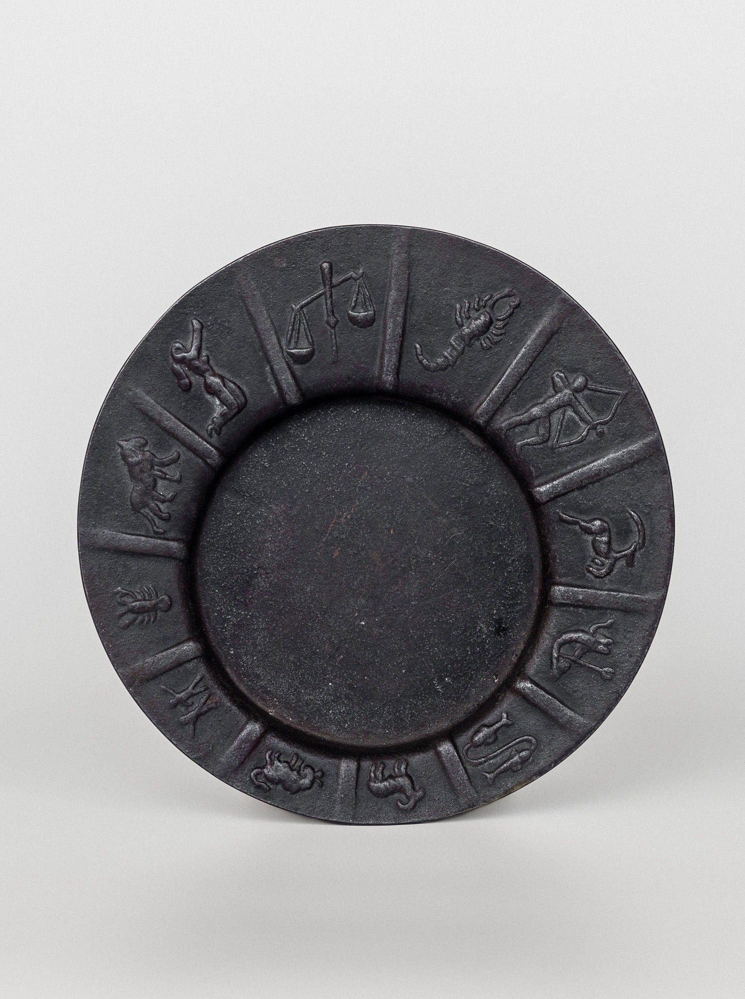 The "Art Deco Zodiac Signs Decorative Dish" by Spigel is a round, dark-colored plate featuring the 12 astrological zodiac symbols embossed along the rim. This Scandinavian vintage design showcases equally spaced symbols starting from Aries and ending with Pisces, arranged clockwise around the center of the dish.