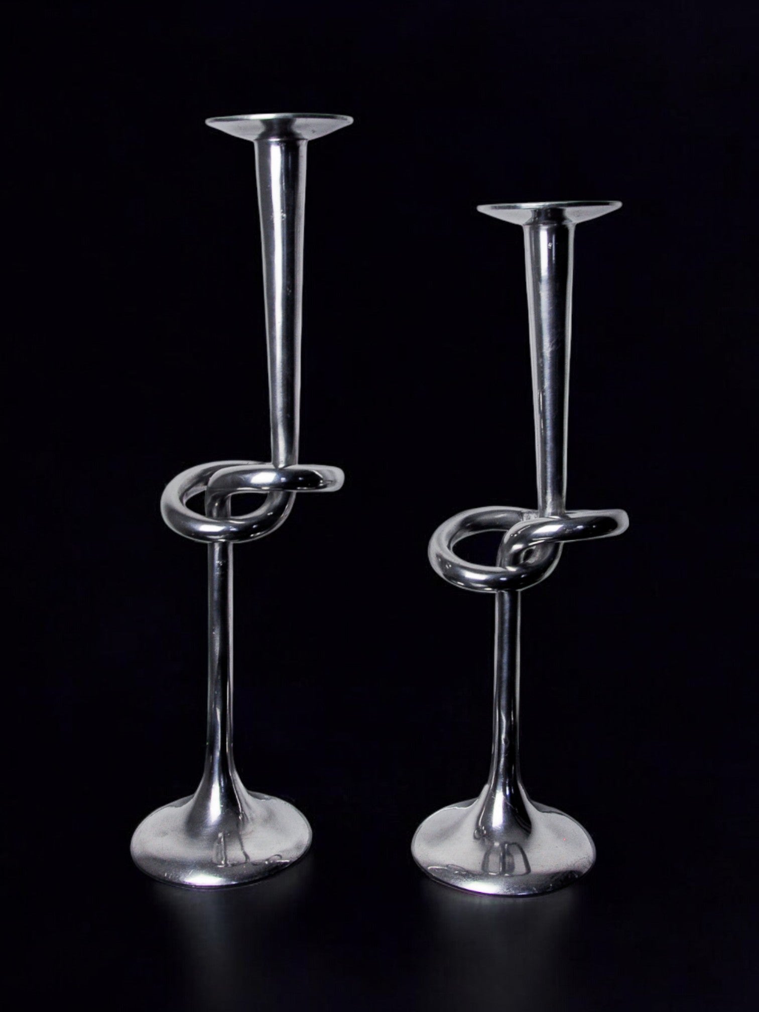 A pair of XL aluminum knotted candlesticks by Malamar Studio, Italy, 1980, feature elegant twisted knot designs along tall stems and shine beautifully against a black background.