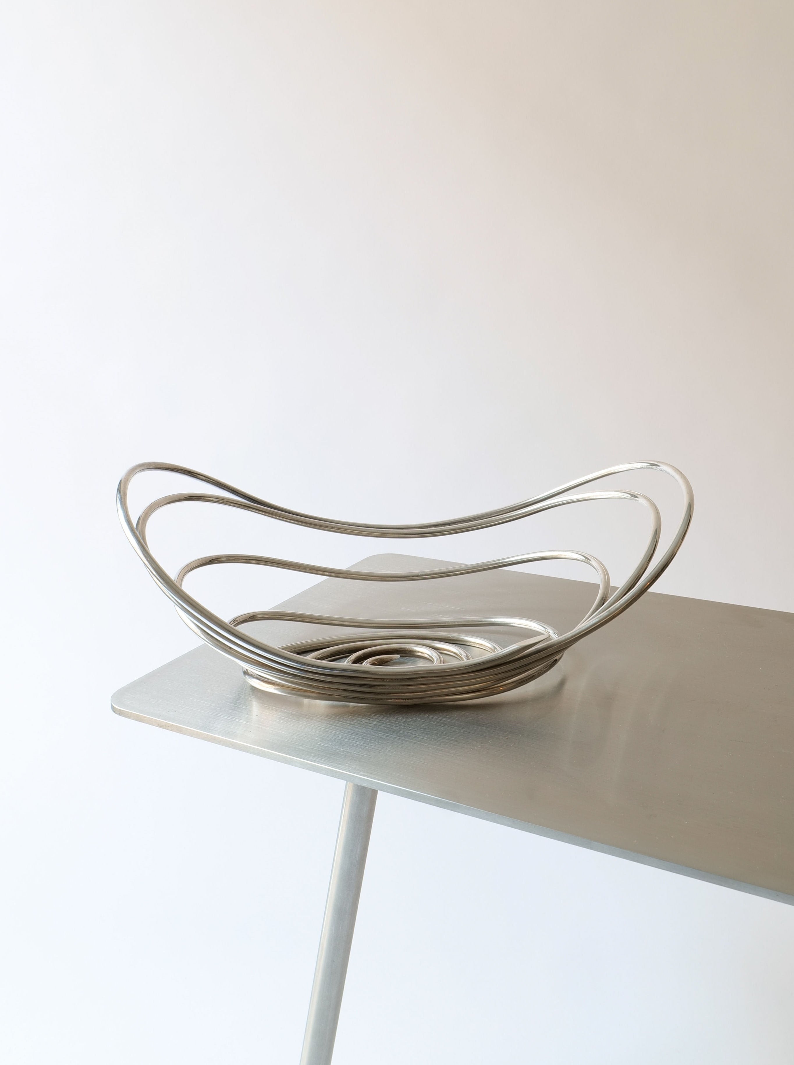 A minimalist metallic fruit bowl, Centrepiece Lino Sabattini 1970s by Collection apart, designed by an Italian designer, with a sleek, open spiral design sits on a modern, light-colored metal table with thin legs. The plain and light background emphasizes the contemporary aesthetic of this highly collectible modernist centerpiece.
