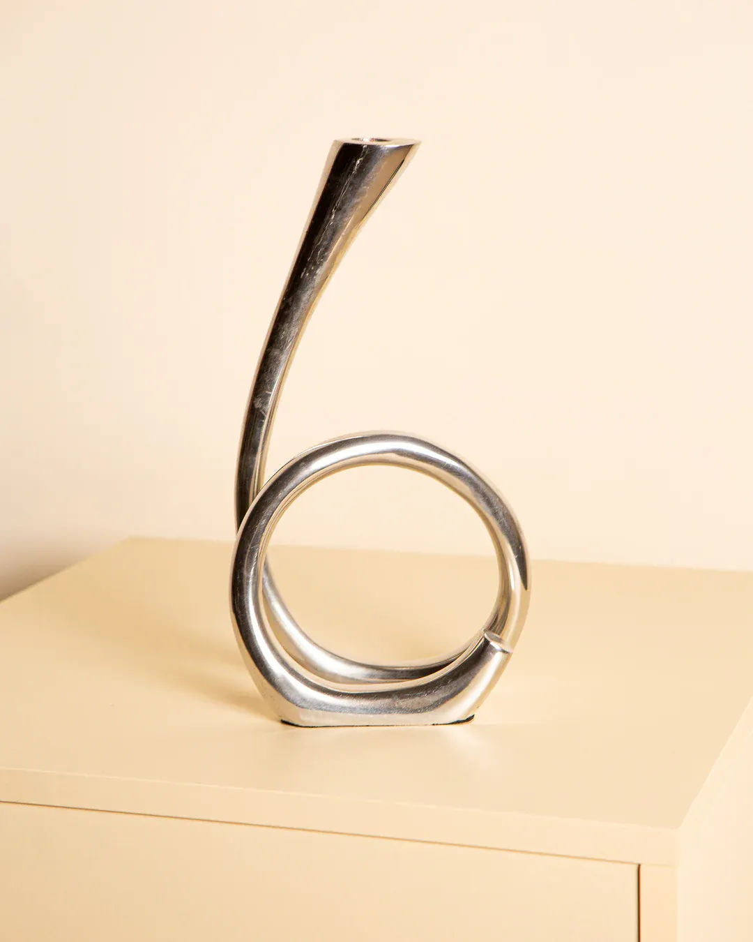 The Treaptyque Cast Aluminum Candleholder 80's, with its sleek modern looped design, rests on a light surface. This minimalist piece resembles a twisted loop with an upward elongated end, embodying the elegance of southern Italy craftsmanship against a plain beige background.