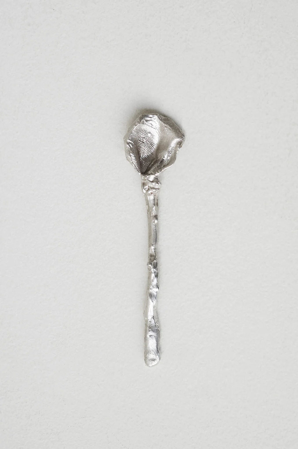 The L32 - Spoon in Silver by Completedworks is a uniquely designed, handcrafted piece with a textured silver finish, featuring an irregular bowl and uneven handle. It showcases UK craftsmanship against a plain, light background.
