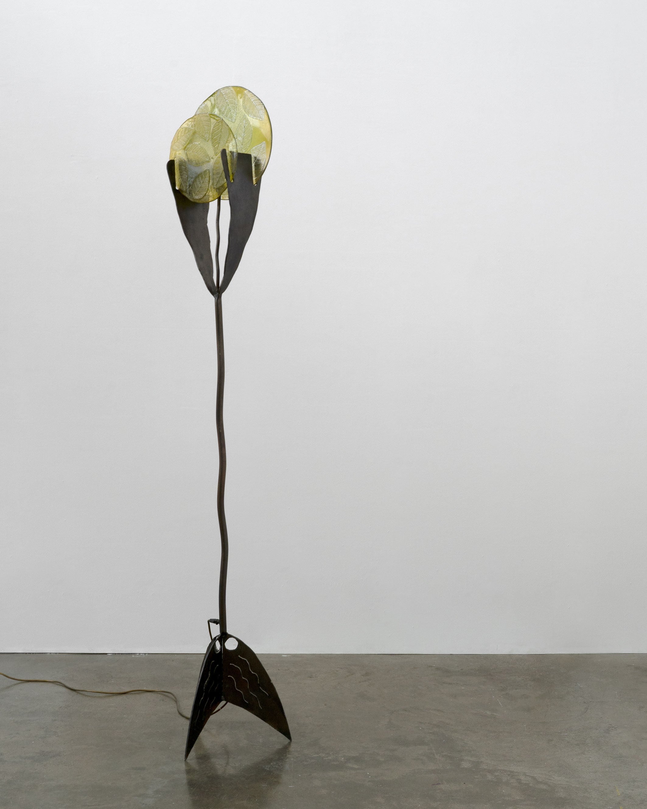 The Brutalist Wrought Iron and Cast Glass Lamp by Tom Bogle (Norway, c.1970) features a wavy black stand, a yellow leaf-inspired shade, and a geometric wrought iron base, set against a white wall and concrete floor.