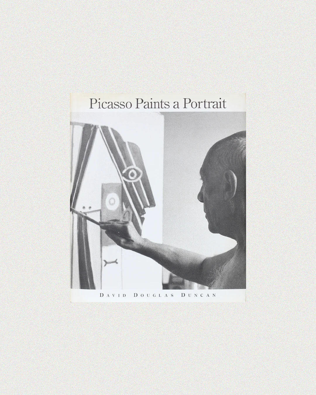 The cover of the book "Picasso Paints a Portrait" from Boga Avante Shop features a striking black-and-white photo of Pablo Picasso holding a brush, painting an abstract eye on canvas.