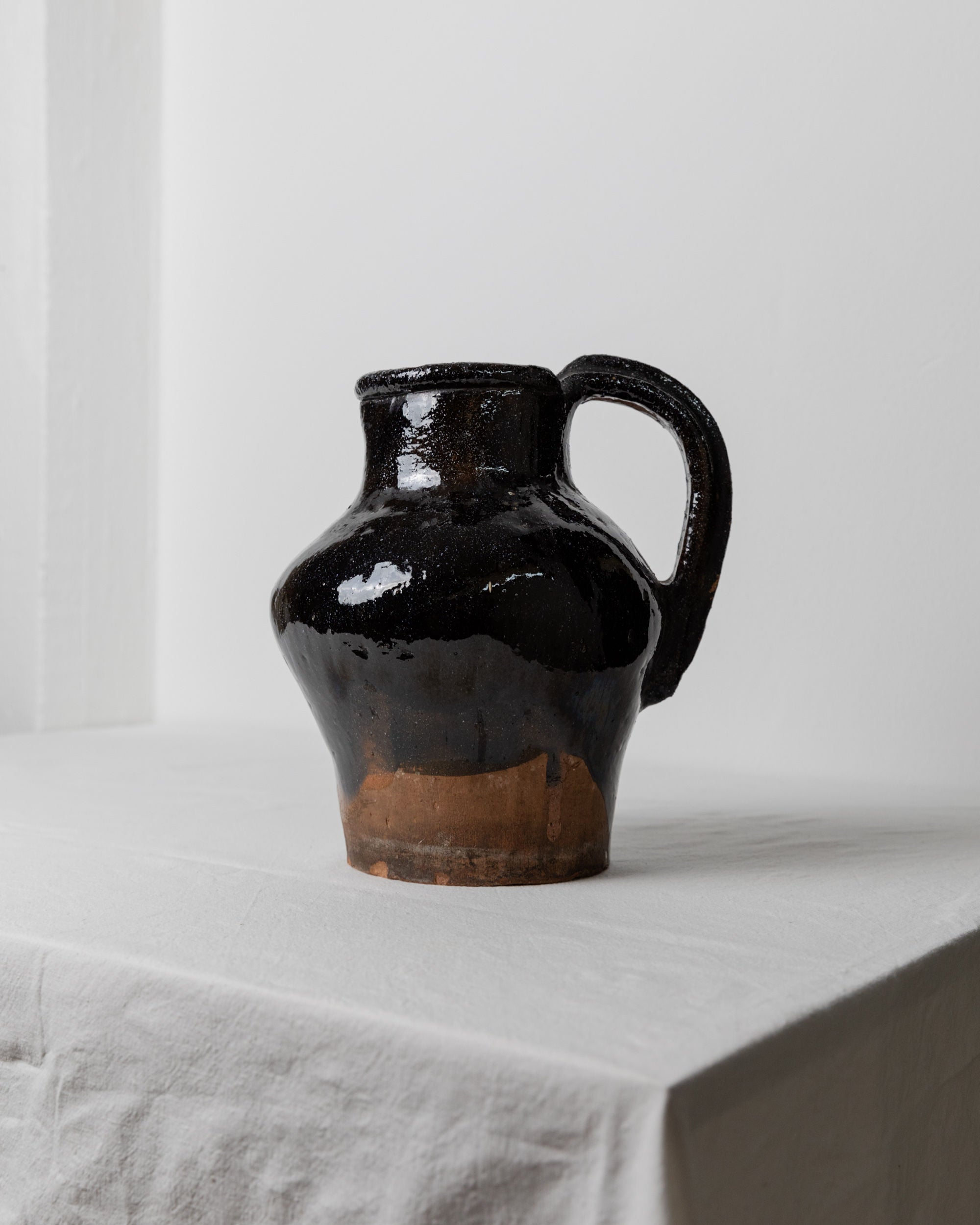 A dark, glossy Antique Terracotta Jug from the 19th century, crafted by Bottega Jacobs in Italy, sits on a white, textured cloth-covered table. The rustic terracotta piece features a wide body and narrow neck, with an unglazed base that reveals natural clay tones. Its vintage design contrasts beautifully against the plain, light-colored wall.